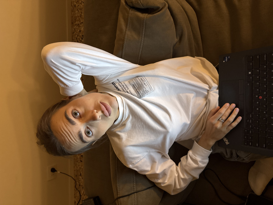 An indoor scene in Venice, Italy, depicts a young adult female looking directly up at the camera while seated or reclining on a dark brown fabric sofa. She has light skin, light-colored eyes, and her hair is pulled back, showing horizontal lines on her forehead. She wears a white long-sleeved mock-neck shirt featuring a rectangular white patch on the left chest with black text, including "CENTURY" and "ITALIAN WHITE MARBLE." Her left arm is bent, with her hand positioned behind her head. Her right hand, wearing a ring on the fourth finger, rests on a black Lenovo ThinkPad laptop placed on her lap. The laptop's keyboard and red trackpoint are visible. Grey trousers are partially visible on the subject. The background features a light-colored wall with a white electrical outlet, from which a black cable extends downwards. Additional dark cables are visible on the sofa near the subject's left side. The scene is lit by warm, directional artificial light.