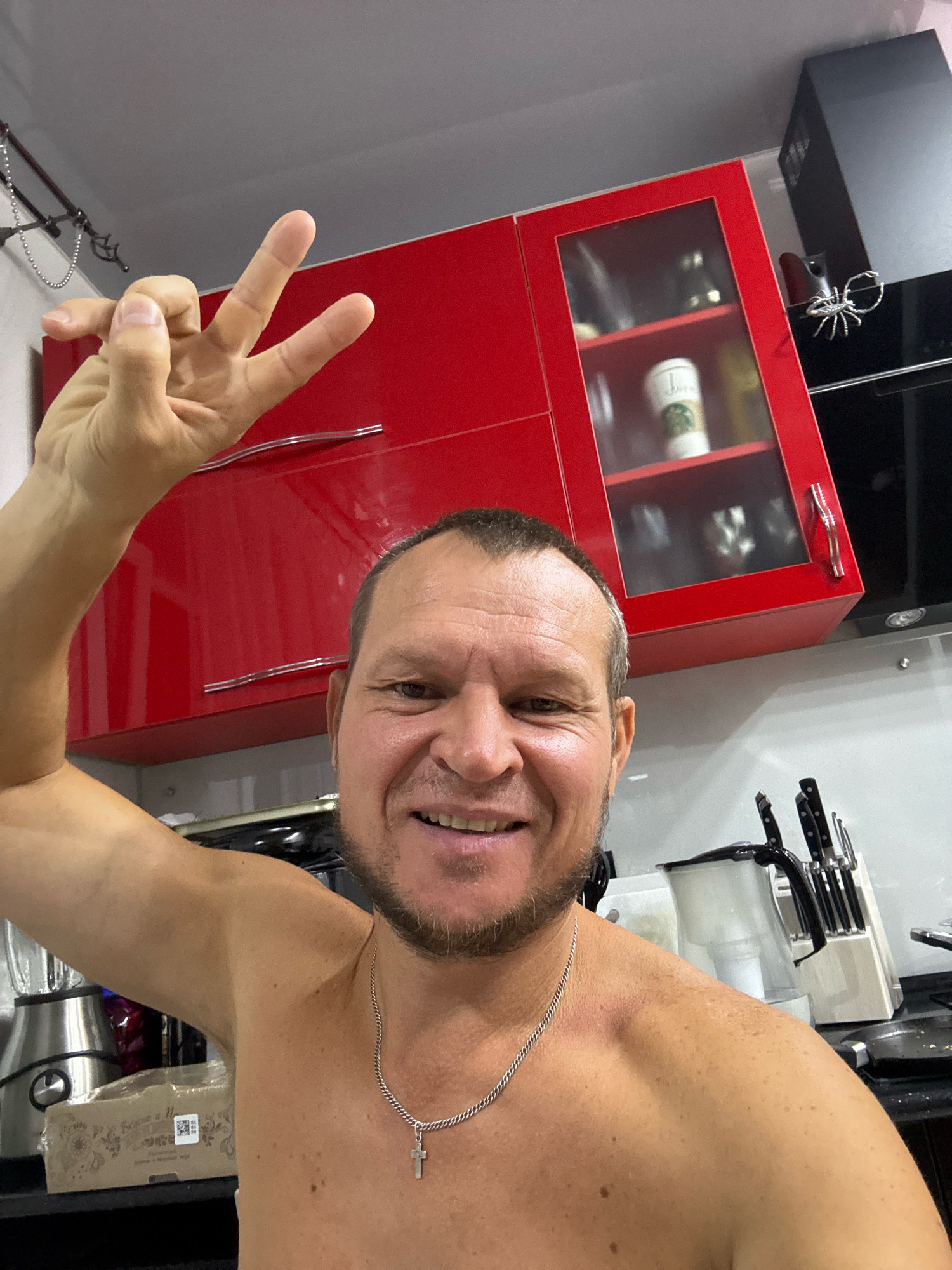 A man, with short brown hair and a beard, smiles at the camera and makes a peace sign with his left hand. He is shirtless and wearing a silver chain with a cross pendant. The man is in a kitchen, with red cabinets visible in the background. A blender is to his left, and a knife block is to his right. The man appears to be in his 40s or 50s. The scene is well-lit and appears to be indoors. The location is not specified within the city of Oskemen, Kazakhstan.