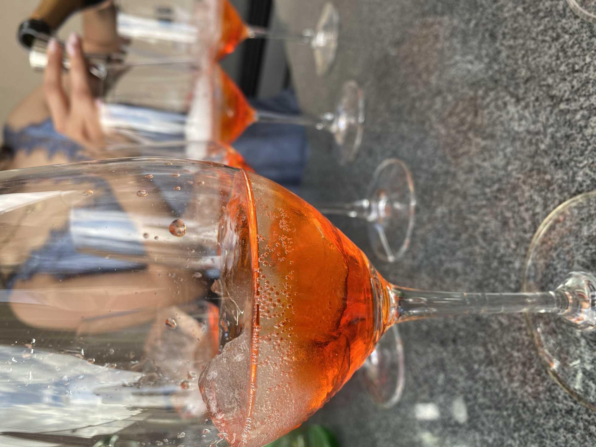 The image shows a close-up view of several wine glasses filled with a light orange-colored liquid, likely a cocktail, containing ice cubes. The glasses are arranged on a gray, speckled surface resembling granite or a similar stone material. In the background, a person appears to be pouring something into one of the glasses, possibly another ingredient for the cocktail. The image's focus is on the texture and appearance of the drinks and the glasses they are served in. The image provides no information about Santa Cruz Tehuixpango, Mexico.