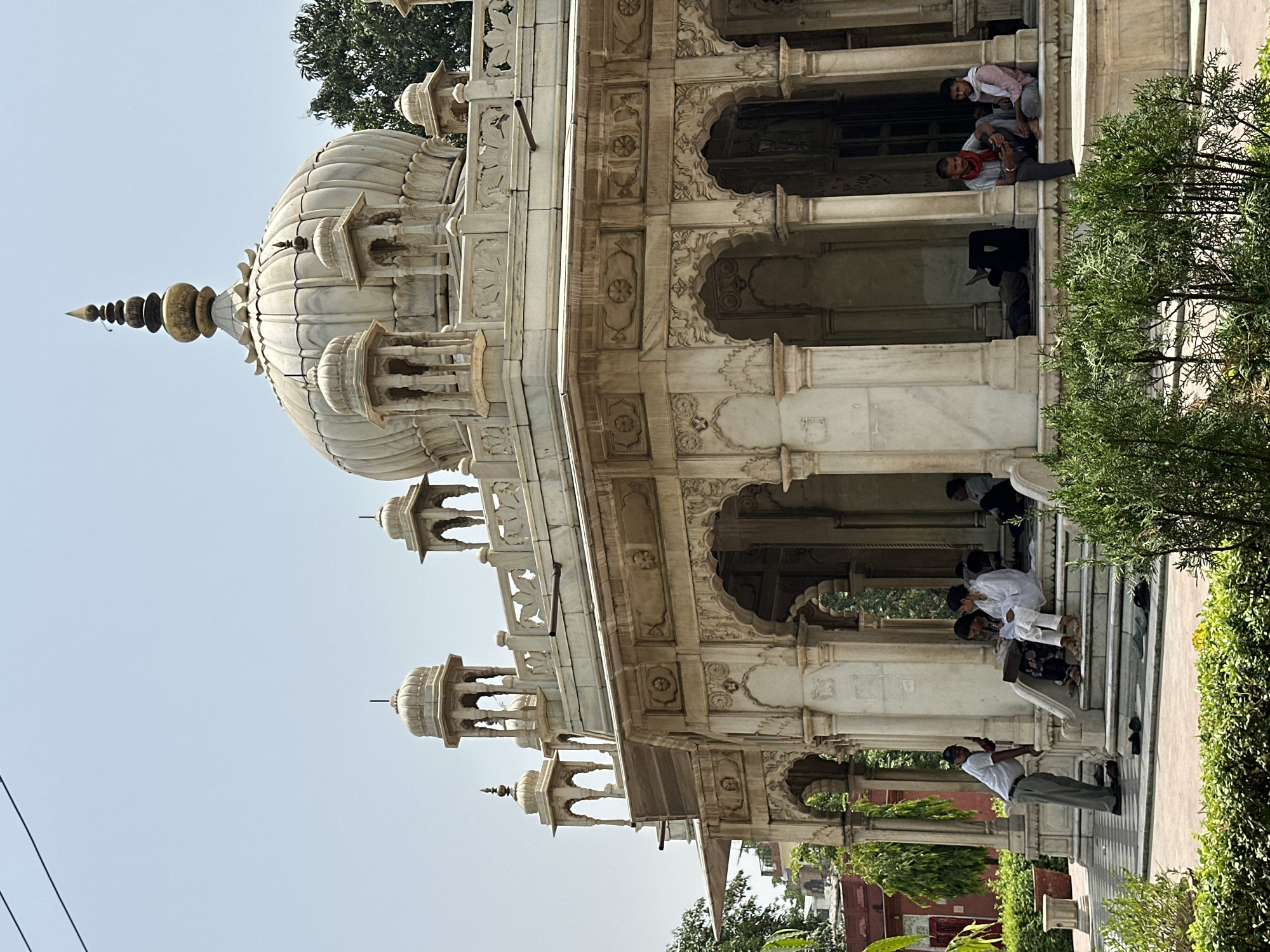The image captures a section of the Alamgir Mosque, located in Varanasi, India. The structure is made of white marble and features Mughal architectural elements, including domes, pillars, arches, and detailed carvings.
Several individuals are present in and around the mosque's courtyard. Some are seated on the steps, while others are standing or interacting within the shaded areas. The architecture and people suggest a place of gathering or resting.
The surroundings include greenery, such as bushes and trees, adding a natural element to the setting. The overall scene presents a blend of historical architecture and everyday life in Varanasi.