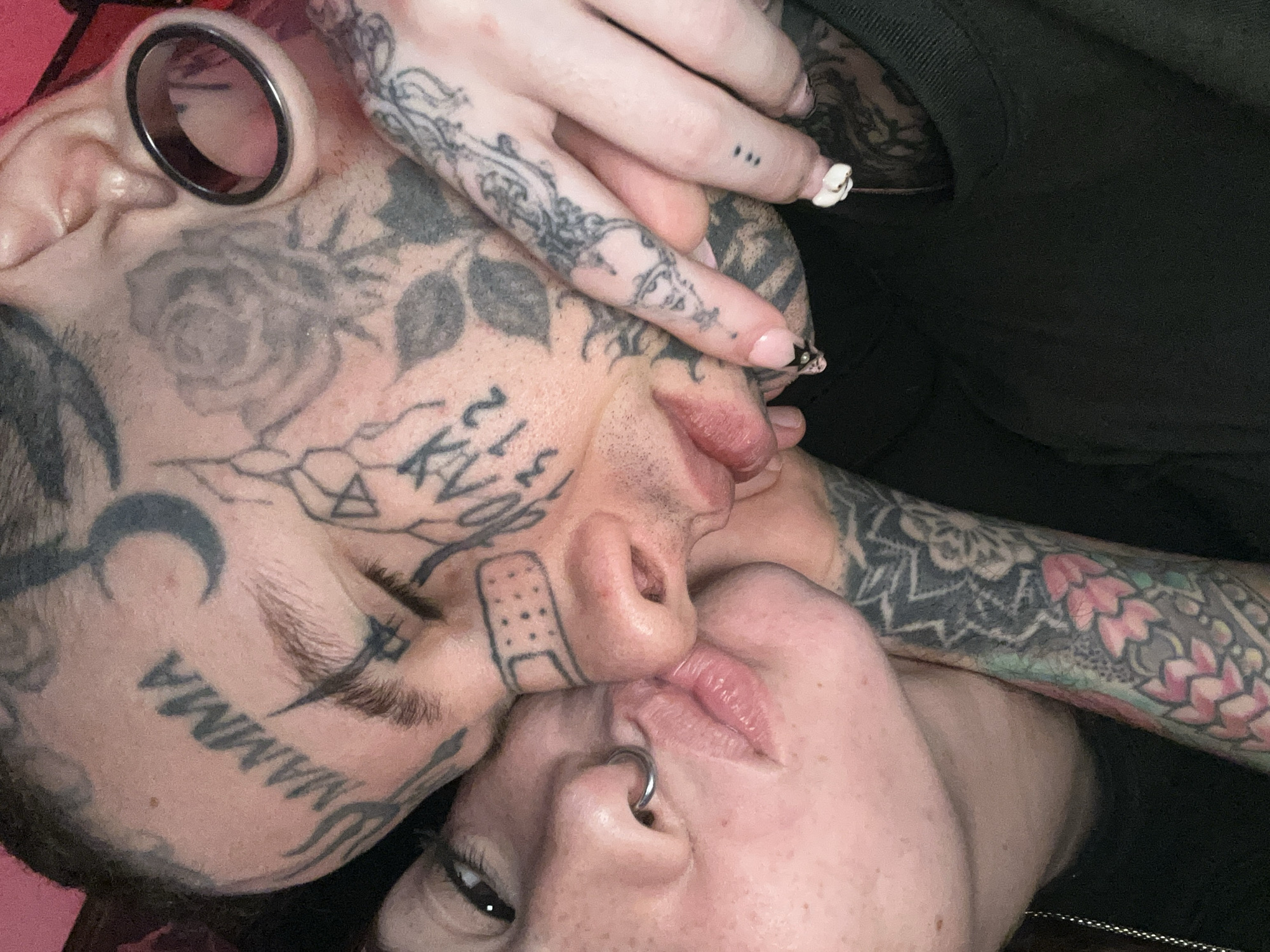 A close-up, top-down shot captures an intimate moment between two heavily tattooed and pierced individuals, likely a couple, in Buenos Aires, Argentina. Their faces are pressed together, with lips touching or nearly touching, conveying affection.
The person on the left (or upper part of the image) is extensively tattooed on the face and neck. Visible tattoos include a large rose on the cheek, the word "MAMMA" in bold uppercase letters along the jawline, a crescent moon, and various geometric and abstract patterns. A distinctive tattoo resembling a band-aid is on the bridge of their nose. They wear a large, black-rimmed ear tunnel or plug.
The second individual, positioned on the right (or lower part), has a septum nose piercing. Their arm, visible in the lower right, is covered with vibrant, intricate tattoos featuring floral and mandala-like designs. A hand, likely theirs, gently cups the first person's head, displaying light pink nail polish. One nail features a small metallic stud, while another has delicate white 3D nail art. Fine-line finger tattoos are also visible, including three small dots on one finger.
The scene is tightly cropped, focusing on the emotional connection and the detailed body art. The background consists of dark fabric and a hint of a pink/red surface, suggesting an indoor, private setting.