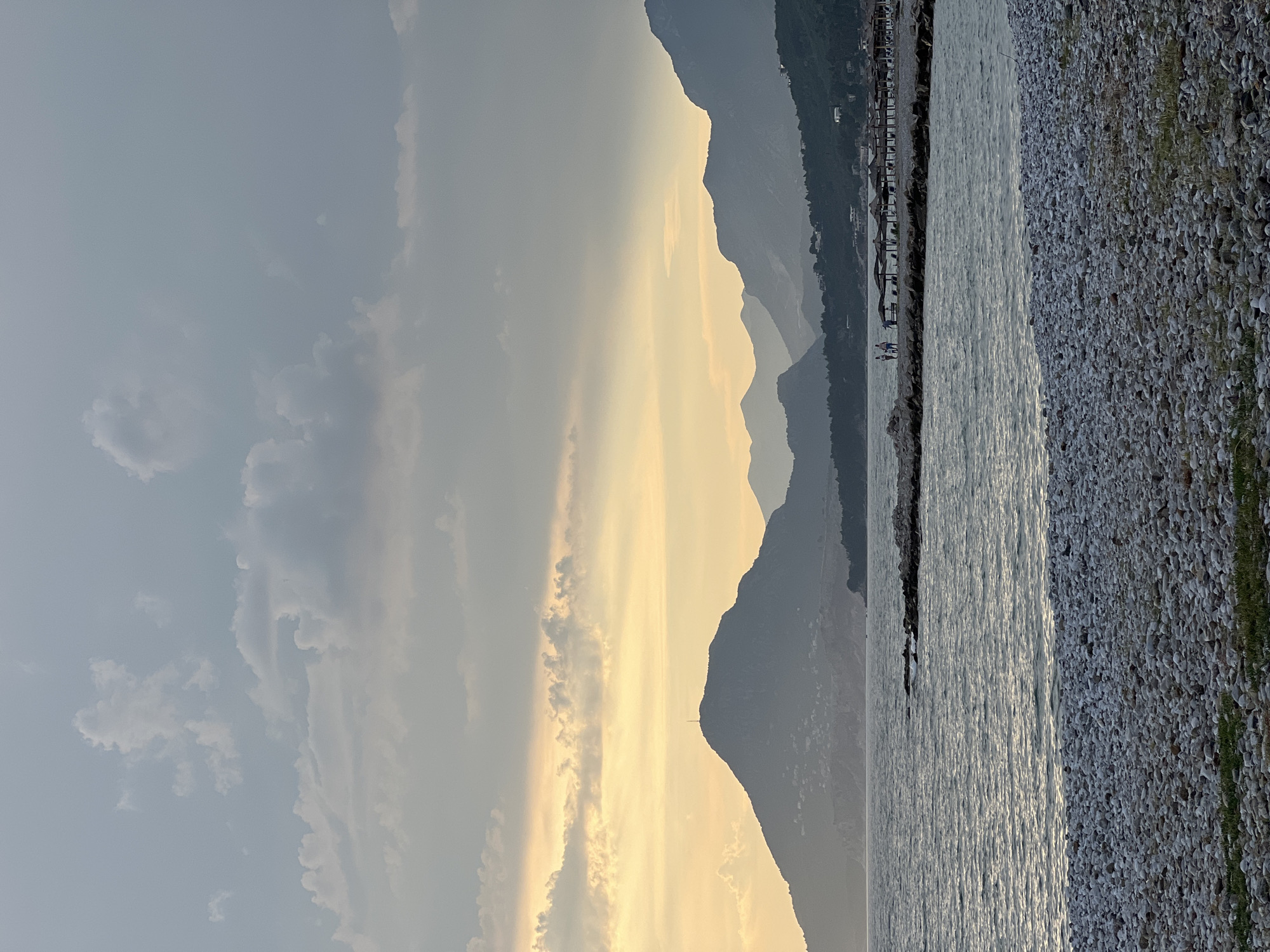 The image depicts a serene coastal scene in Bar, Montenegro, likely during twilight. In the foreground, a pebbled beach stretches across the frame, with a gentle slope leading to the calm sea. The water exhibits a rippling texture, reflecting the subdued light of the sky. Further out, a rocky outcrop juts into the sea, adorned with a few scattered beach umbrellas and a small cluster of people, suggesting a quiet recreational area. Beyond the water, a series of majestic, silhouetted mountains form the backdrop. The largest mountain in the mid-ground is crowned with a prominent antenna, its slopes dotted with faint lights indicating a settlement. The sky above is a canvas of soft hues, with streaks of pale yellow and orange from the setting sun mingling with cooler grey and blue tones, interspersed with scattered clouds. The overall atmosphere is tranquil and picturesque, capturing the natural beauty of the Montenegrin coastline.
