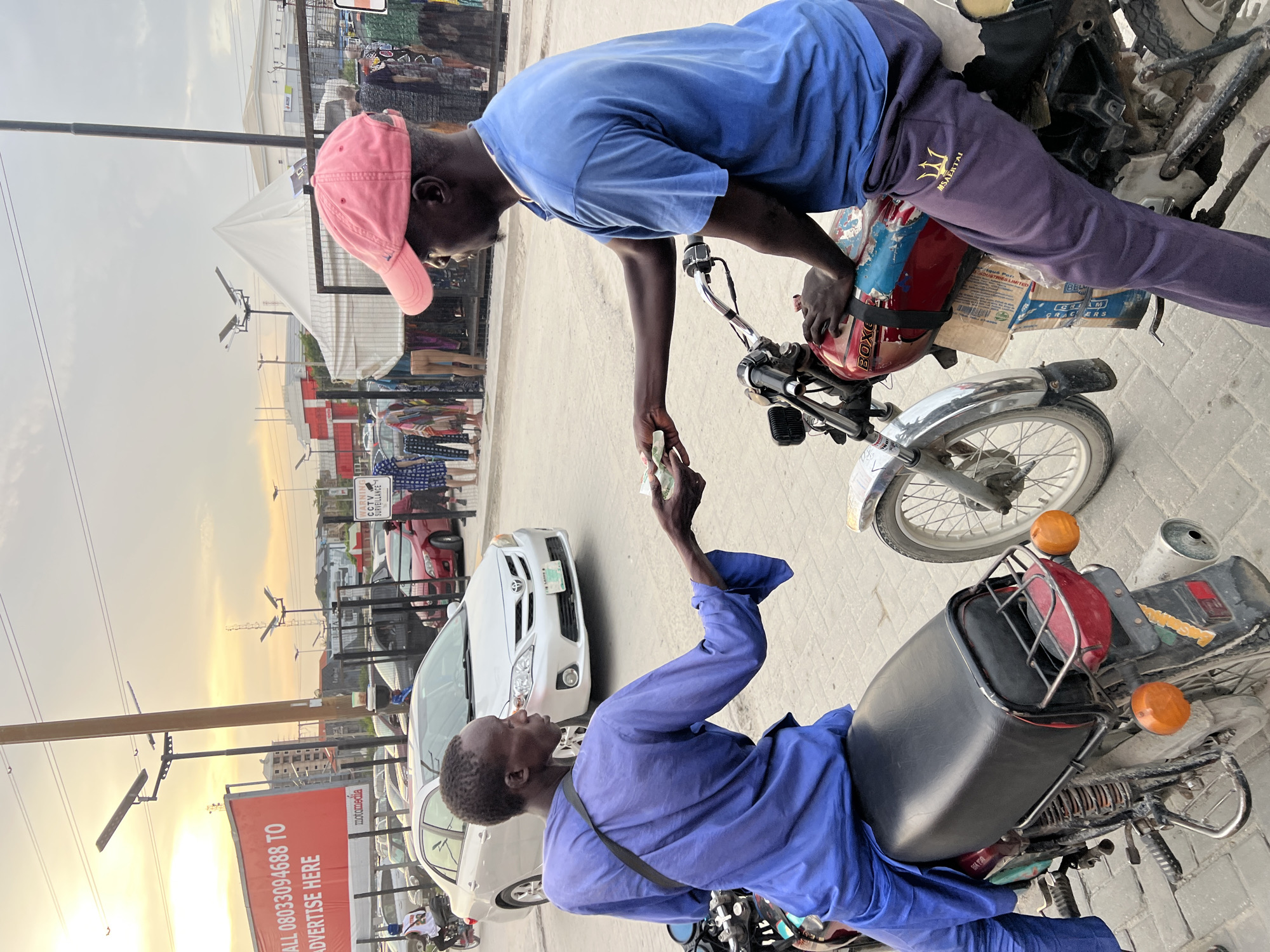 The image captures a street transaction between two men in Igboefon, Nigeria, set during what appears to be late afternoon or early evening with a warm, low sun visible in the background.
The primary subjects are two Black men. One, wearing a blue short-sleeved t-shirt, dark purple sweatpants with a trident-like logo and text (possibly "MSAERTAL"), and a pink baseball cap, stands on the left. He is leaning towards the right, extending his hand to offer or receive something, likely money, to the second man. This second man is seated on a motorcycle on the right, wearing a blue long-sleeved shirt and dark pants, and is reaching out to complete the exchange.
Both men are positioned near motorcycles. The man in the pink cap stands beside a motorcycle with a red helmet ("BOXE" visible) resting on its tank, and a cardboard box labeled "BELLA CREAM CRACKERS" is secured to its rear. The seated man is on another motorcycle, which has a black passenger seat and orange turn signals.
In the background, various street elements are visible. These include other cars, such as a white Toyota Corolla and a red car, parked along the road. Street vendor stalls or market setups can be seen further back. Utility poles with solar-powered lights line the street. A prominent red billboard is visible on the left, displaying white text: "CALL 08033094688 TO ADVERTISE HERE" and "motormedia". Another small sign warns "CCTV SURVEILLANCE". The sky, especially towards the horizon, shows a golden hue from the setting sun, with some clouds present.