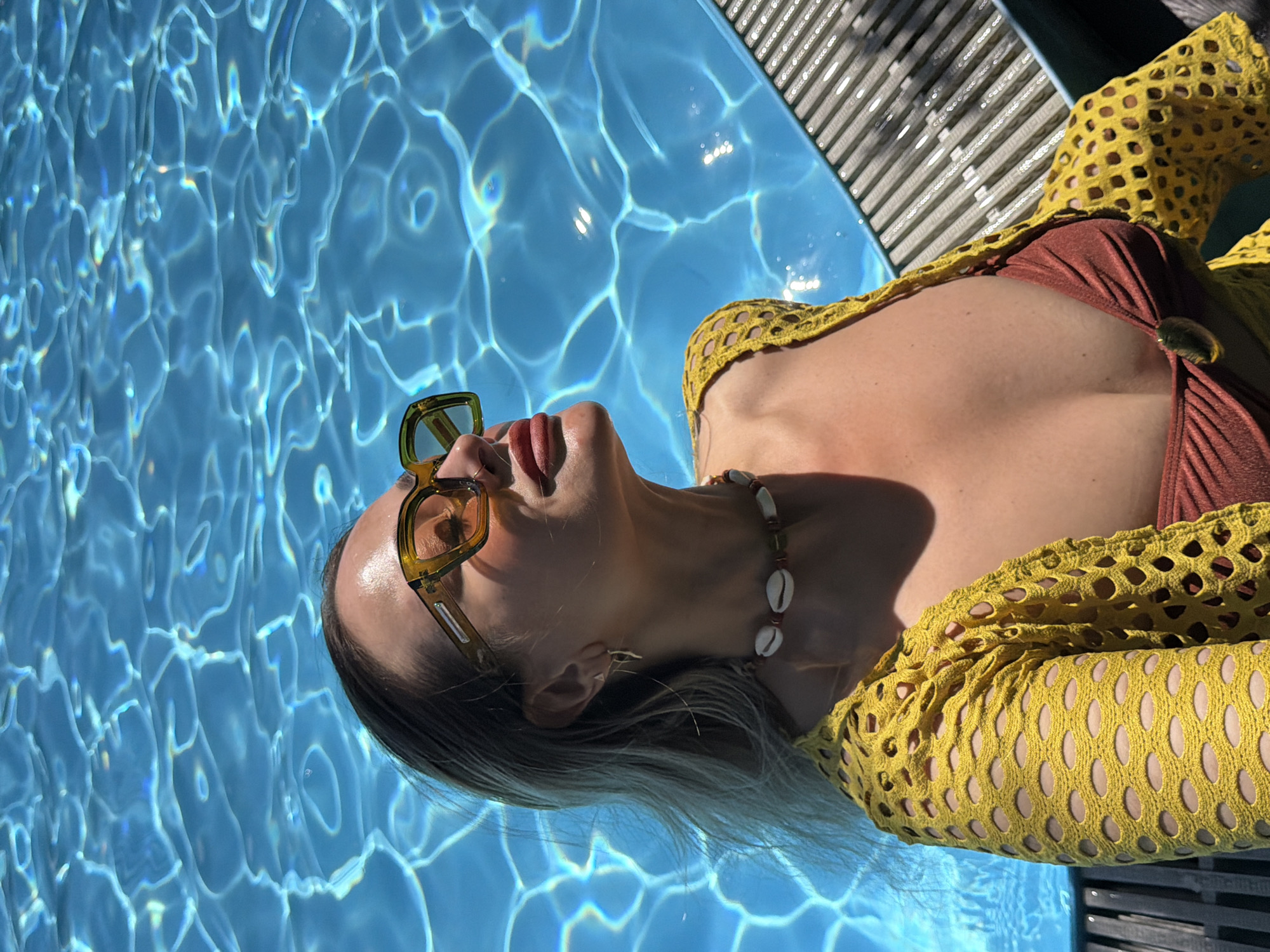 This media file captures a person, likely a woman, relaxing by a swimming pool on a sunny day in Krasnoyarsk, Russia. The individual is depicted lying on their back, with their head tilted upwards. They are wearing a brown bikini top and a distinctive yellow, open-knit or crocheted cover-up featuring a geometric hole pattern. Olive-green framed sunglasses cover their eyes, and a beaded necklace adorned with small white cowrie shells is visible around their neck. Long, light-colored hair trails into the clear blue water of the pool, which reflects shimmering patterns of sunlight. A dark-colored pool grating or edge is partially visible along the right and top side of the frame. The strong, natural sunlight casts distinct shadows, indicating a bright, clear day. The person appears relaxed. There is no visible text in the image.