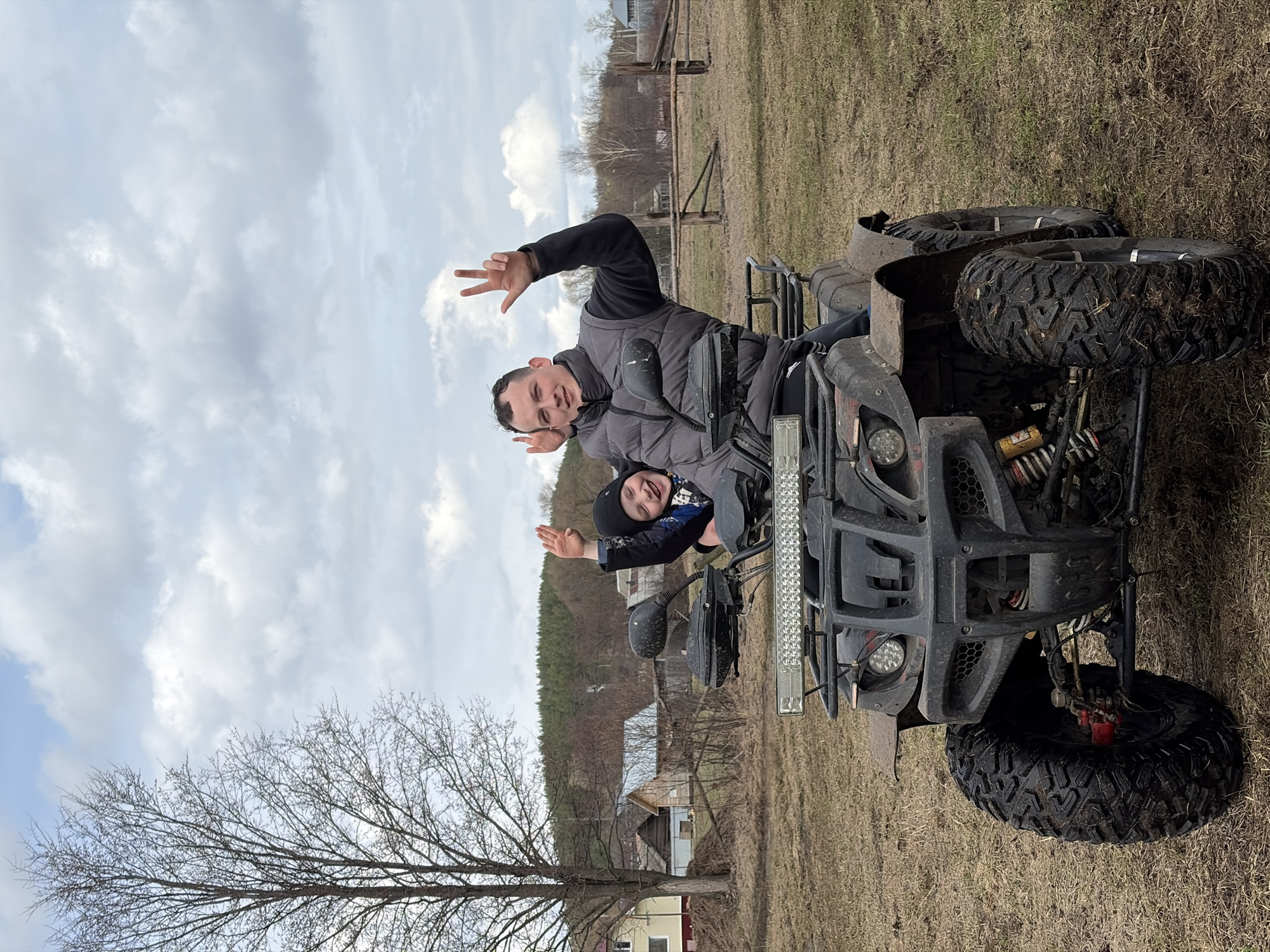 The image shows two people on an ATV in a rural setting, likely in Кемуль, Russia. An adult male is seated on the ATV, gesturing with his hand. A child is standing in front of him. The ATV is in a field with sparse vegetation. There are trees and buildings in the background. The sky is overcast.