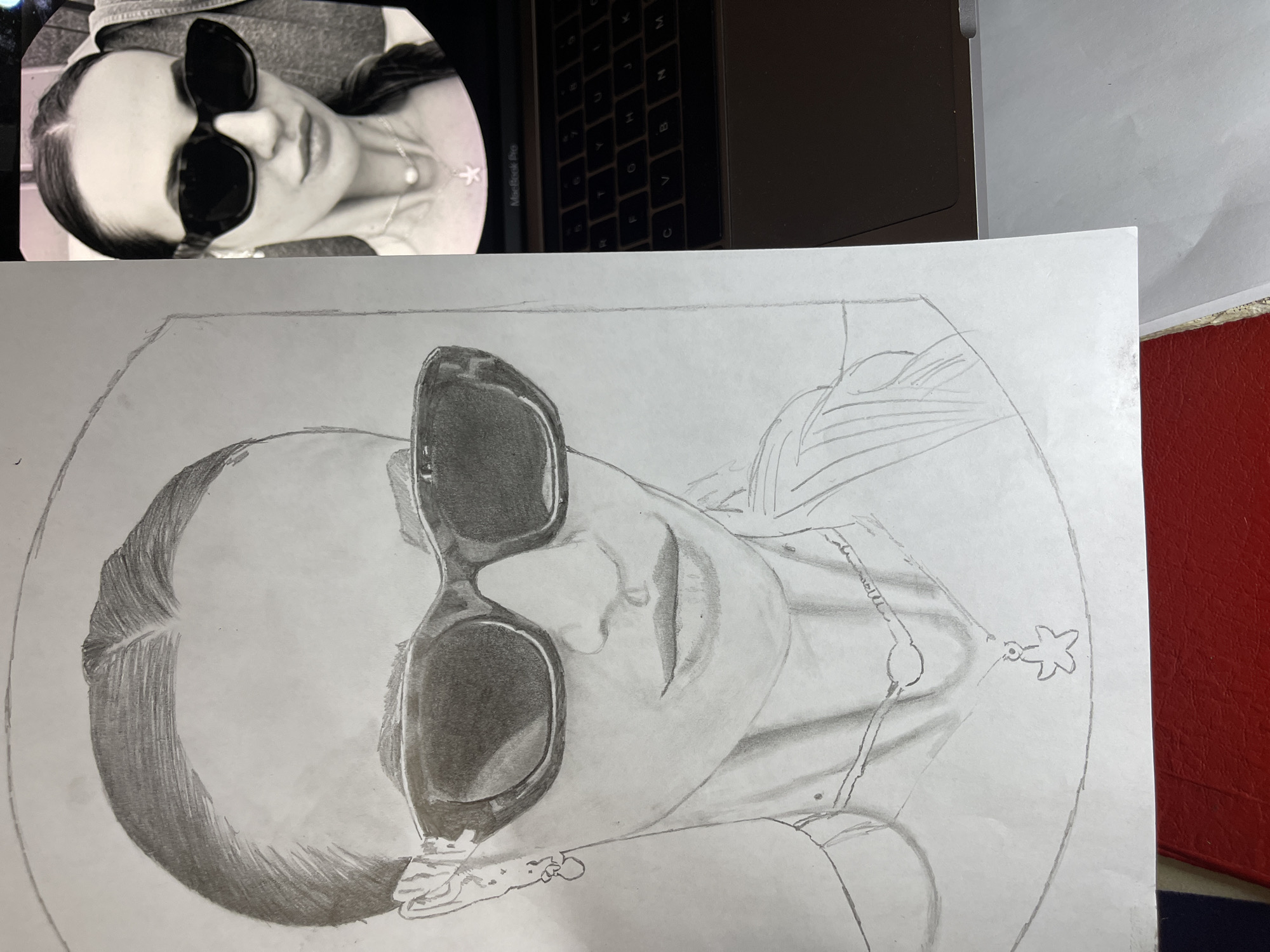 The image shows a partially completed pencil drawing of a woman wearing sunglasses and a necklace with a starfish pendant. The drawing is on a white sheet of paper placed next to a laptop. The laptop shows a photographic reference of the woman being drawn, and part of the laptop keyboard is visible. The drawing is oriented similarly to the photographic reference, featuring the woman's head and shoulders, with visible shading and line work defining the facial features, hair, and sunglasses. A red surface is also partly visible. The location in Jos, Nigeria is not specified in the image.