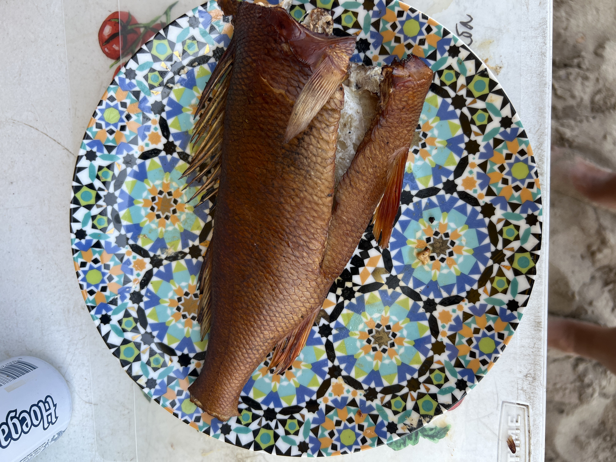 A whole, cooked fish with reddish-brown scales and fins lies on a round, vibrantly patterned plate. The plate features a geometric mosaic design with blue, green, orange, black, and white star-like shapes. A portion of a white can with black lettering is visible in the upper left corner. The plate is resting on a light-colored surface, possibly a table. A faint image of red tomatoes is visible on the surface behind the plate. The background appears to be a sandy or granular texture. There are no people visible in the image. The location is not specified in the image or its metadata.