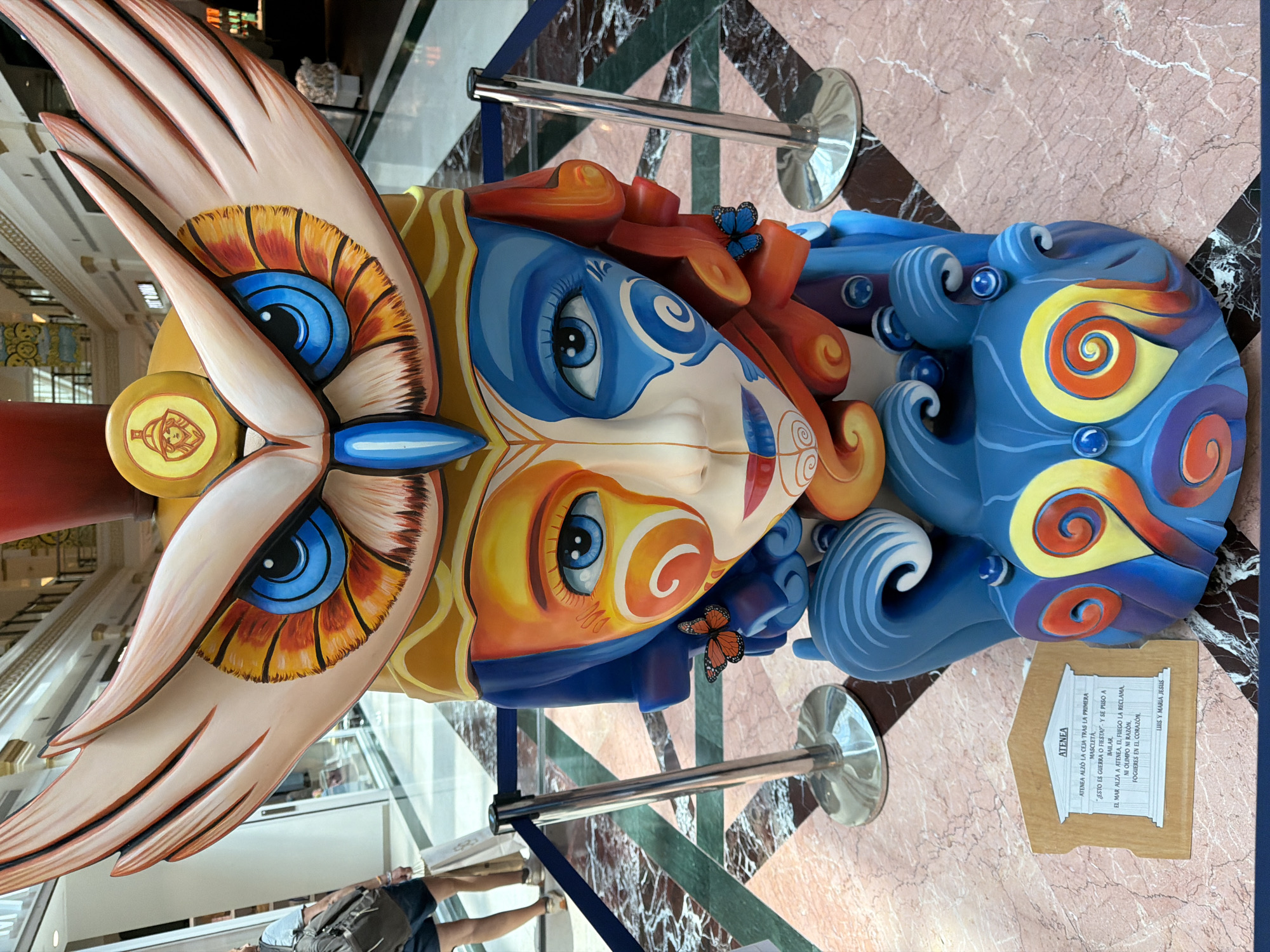 The image displays a large, colorful sculpture in what appears to be an indoor public space, likely a mall or museum, given the polished marble floor, storefronts in the background, and stanchions with velvet ropes. The sculpture is a vibrant, stylized representation of a face with owl-like features. The face is split vertically down the middle, with one side painted in warm orange and yellow tones and the other in cool blue. Large, expressive blue eyes are prominent, framed by feathered owl-like "eyelids" that extend outwards like wings. The sculpture has a distinctive headdress that resembles an owl's head with large wings. The lower part of the sculpture is a flowing, wave-like structure in shades of blue, adorned with colorful swirls and small blue spheres. Two small orange butterflies are affixed to the sculpture's hair-like elements. A small wooden plaque with text is placed at the base of the sculpture. In the background, two people are visible, one walking away from the camera carrying a backpack and a shopping bag, and another standing further back. The lighting is bright and even, suggesting an indoor environment.