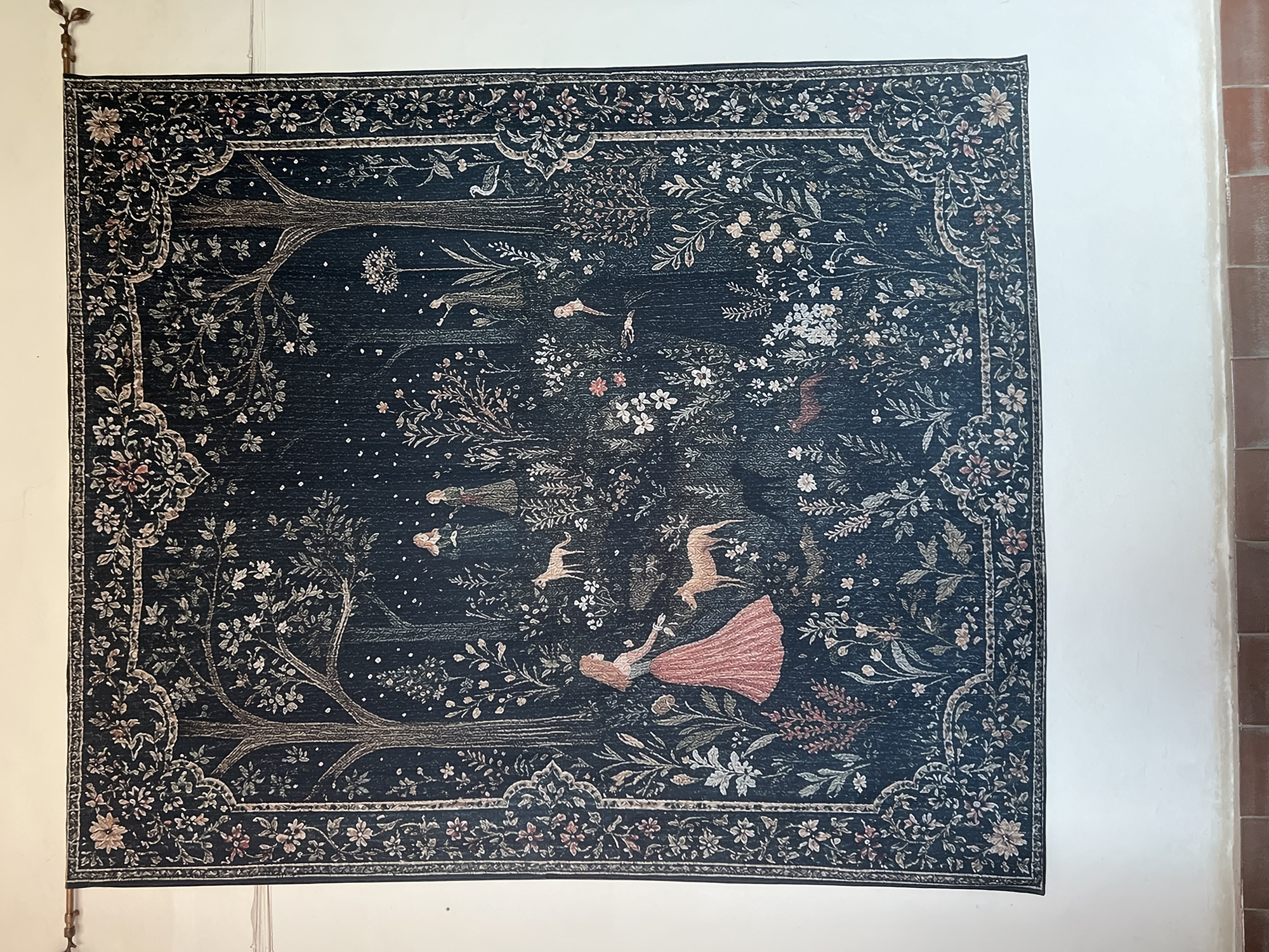 The image shows a dark blue tapestry hanging on a white wall. The tapestry depicts a scene with trees, flowers, and several figures. In the foreground, a woman in a pink dress is interacting with two deer-like animals. Further back, other figures are visible, as well as more trees and plants. The tapestry is framed by an ornate border featuring floral motifs. The wall is plain white, and the floor below is tiled with reddish-brown tiles. The scene is indoors, possibly in a home or museum, in the city of Karpaty, Ukraine.