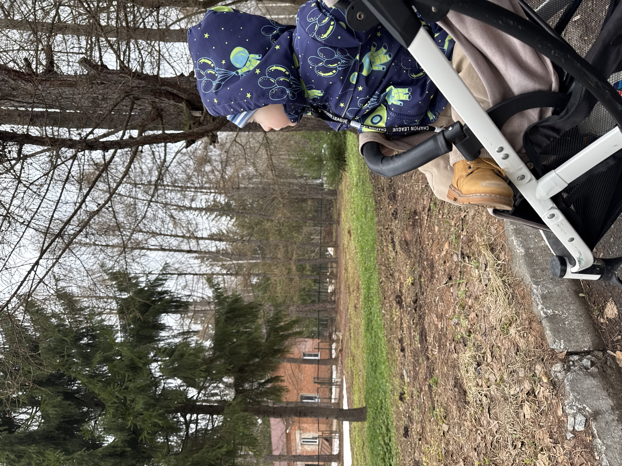 A child in a blue patterned jacket and tan boots is sitting in a stroller outdoors. The background features trees and a building partially visible behind the trees. The setting appears to be a park or wooded area near a building. The location is in Izhevsk, Russia.