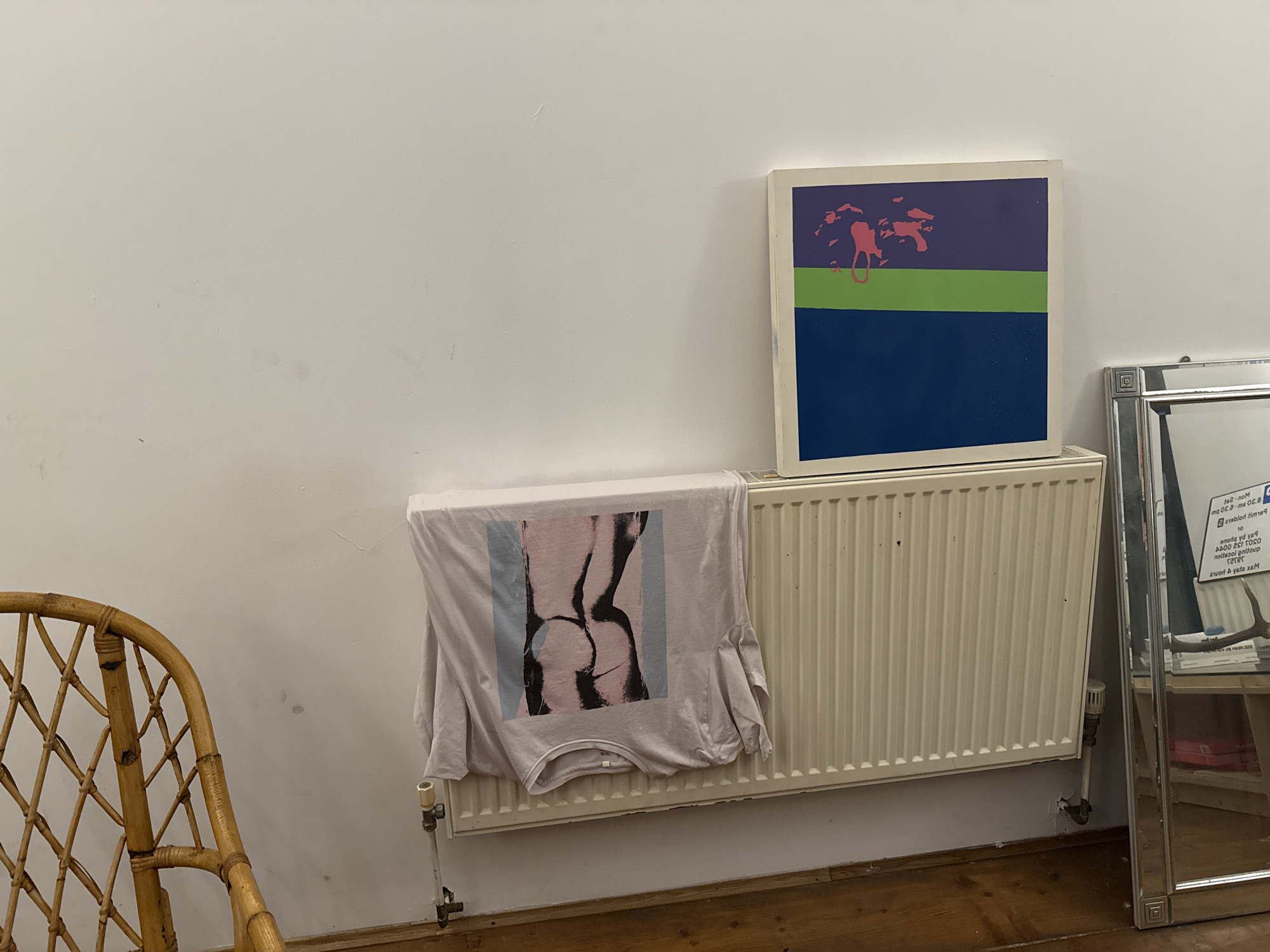 The photograph shows an interior space, likely a room in a house or apartment. The walls are white. A light-colored, woven wicker chair sits on a wooden floor on the left of the image. A white radiator is positioned against the wall. A t-shirt with a graphic image is draped over the radiator. On top of the radiator rests a square painting or print with horizontal blocks of navy, green, and purple, with some pink shapes on the purple section. To the right, a silver-framed mirror leans against the wall. There is text visible on a paper in front of the mirror. The location of this image is not explicitly provided, but based on the details of the text visible and its location in the United Kingdom is the city of London. There are no people visible.