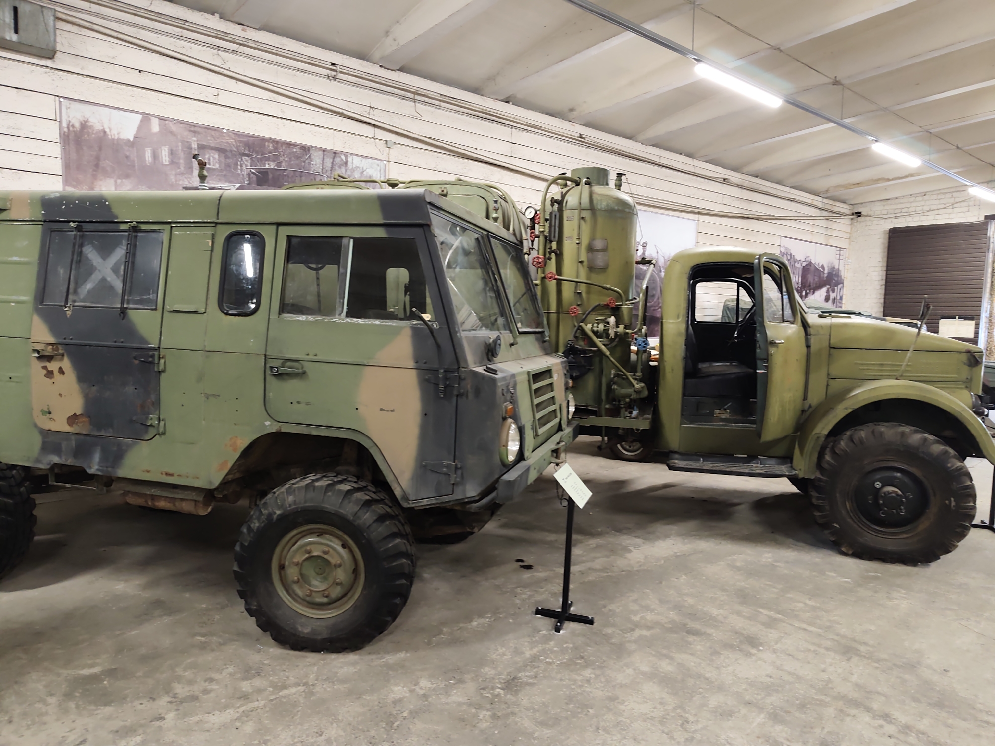 The image depicts two military-style vehicles parked indoors. The vehicle on the left is a green and camouflage-painted truck with a boxy cargo area and large tires. It has a closed cab with several windows, some of which are covered with white tape in an 'X' pattern. The vehicle on the right is an older-style olive green truck with an open driver's side door, revealing the interior. Attached to the rear of the cab on the right vehicle is a large, cylindrical tank with various pipes and valves. The vehicles are parked on a concrete floor in what appears to be a museum or exhibition hall, as indicated by the industrial lighting fixtures on the ceiling and large pictorial backdrops on the wall. The backdrop on the left shows a black and white image of buildings and trees. A black metal stand with a sign is positioned in front of the left vehicle. The environment is clean and well-lit. This scene is located at the Viimsi Open Air Museum in Viimsi, Estonia.