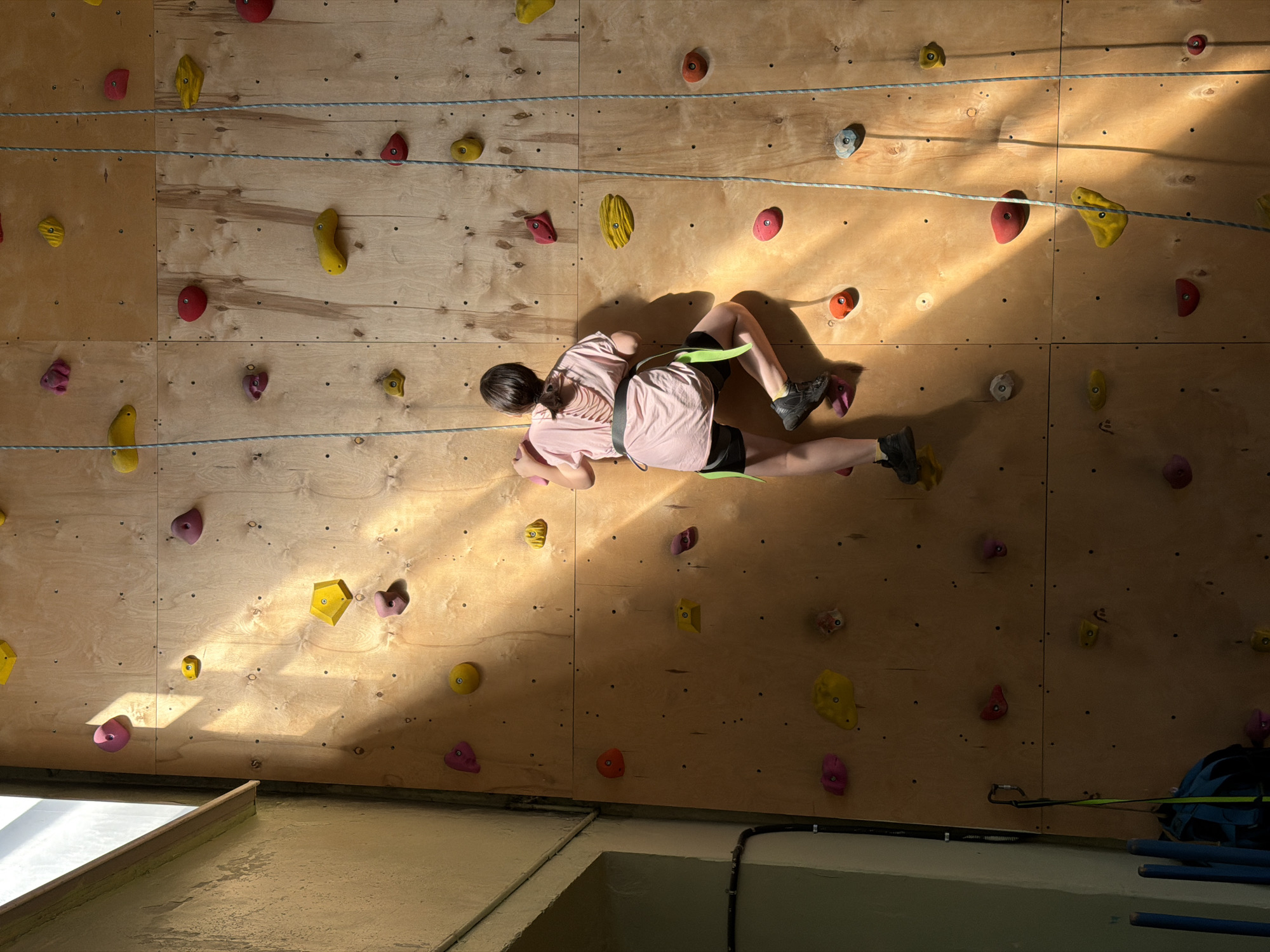 A girl is climbing on an indoor rock climbing wall. The wall is made of wood and has a variety of colorful climbing holds attached to it. The girl is wearing a pink shirt, black shorts, and climbing shoes. She is also wearing a harness and is attached to a rope. The location is an indoor climbing gym; the city, Izhevsk, Russia, is not evident in the image.