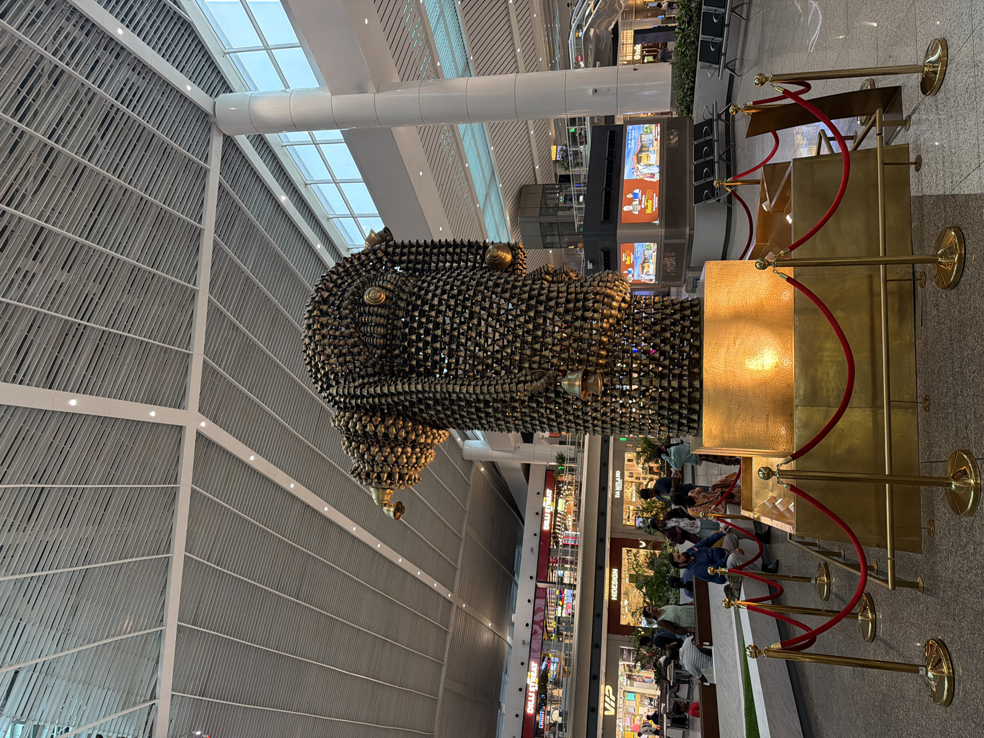 The image depicts the interior of Indira Gandhi International Airport in New Delhi, India. A large, intricate sculpture made of numerous brass bells stands prominently in the foreground. The sculpture is gold-colored and appears to be illuminated from within its base. It is surrounded by a golden barrier with red velvet ropes, guiding visitors and preventing close contact. In the background, there are shops and businesses, including "DILLI STREET," "VIP," "HIDESIGN," and "DA MILANO." People are visible walking around the airport, with some seated on benches or browsing in the shops. The airport features a modern, spacious design with a high, slatted ceiling and large windows that allow natural light to enter. Escalators and seating areas are also visible in the background. The overall scene is one of a busy and well-maintained international airport terminal.