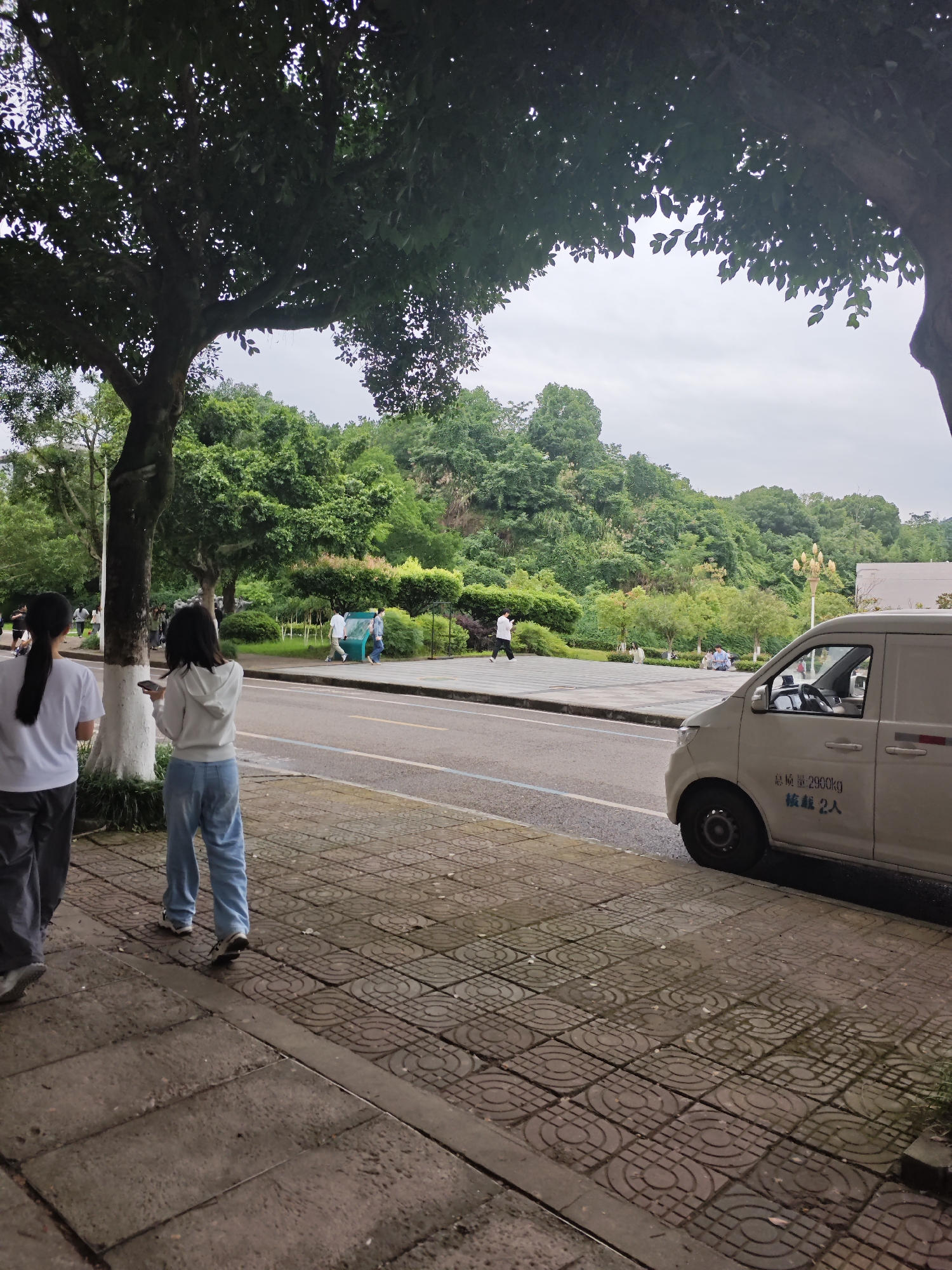 The image depicts a street scene in the Chongqing High-tech Zone, China, on an overcast day. In the foreground, two young women are walking on a sidewalk made of patterned concrete tiles. The woman on the left wears a white t-shirt and dark pants, while the woman on the right wears a white hoodie and light blue jeans, and is holding a smartphone. A large tree with dense foliage shades part of the sidewalk.
Beyond the sidewalk is a road with a light-colored van parked on the right side. The van has Chinese text on its side indicating a weight limit of 2900kg and a capacity of 2 people. In the background, across the road, is a public plaza with paved flooring. Several people are visible in the plaza, including a man walking and a group of people sitting. The plaza is bordered by manicured green spaces with trees and shrubs. Beyond the plaza, a densely forested hillside rises, characteristic of the natural landscape in the region. The overall atmosphere is calm and ordinary, suggesting a typical day in this urban park-like setting.