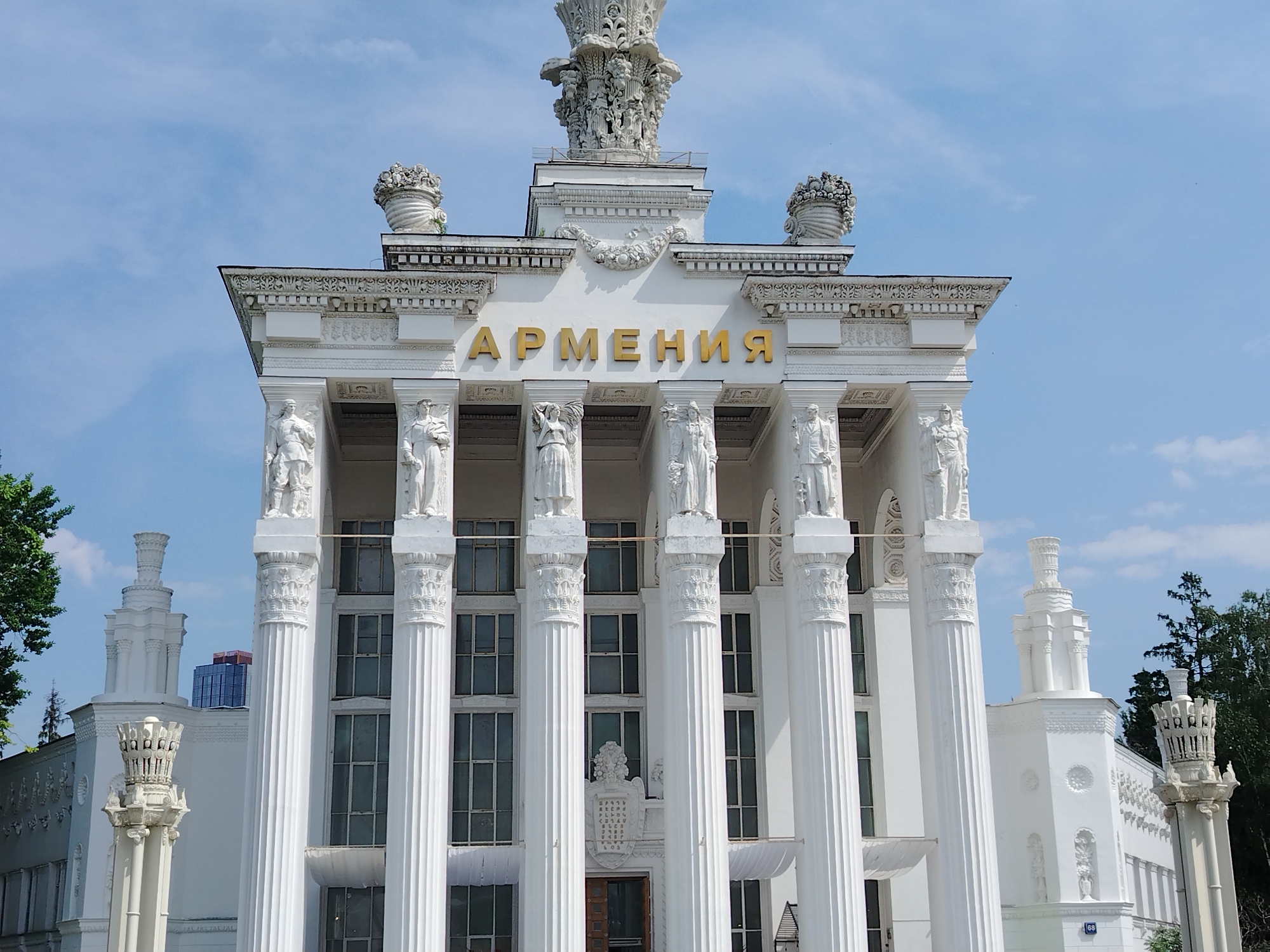 The image shows the Armenia Pavilion at VDNKh (Exhibition of Achievements of National Economy) in Moscow, Russia. The pavilion is a white, neoclassical structure with a large "АРМЕНИЯ" (Armenia) sign in gold letters across the top. It features a row of columns, each topped with a statue. The entrance has an ornate pediment. The sky is blue with some clouds. Trees are visible on either side of the pavilion. Other buildings can be seen partially obscured behind the pavilion.