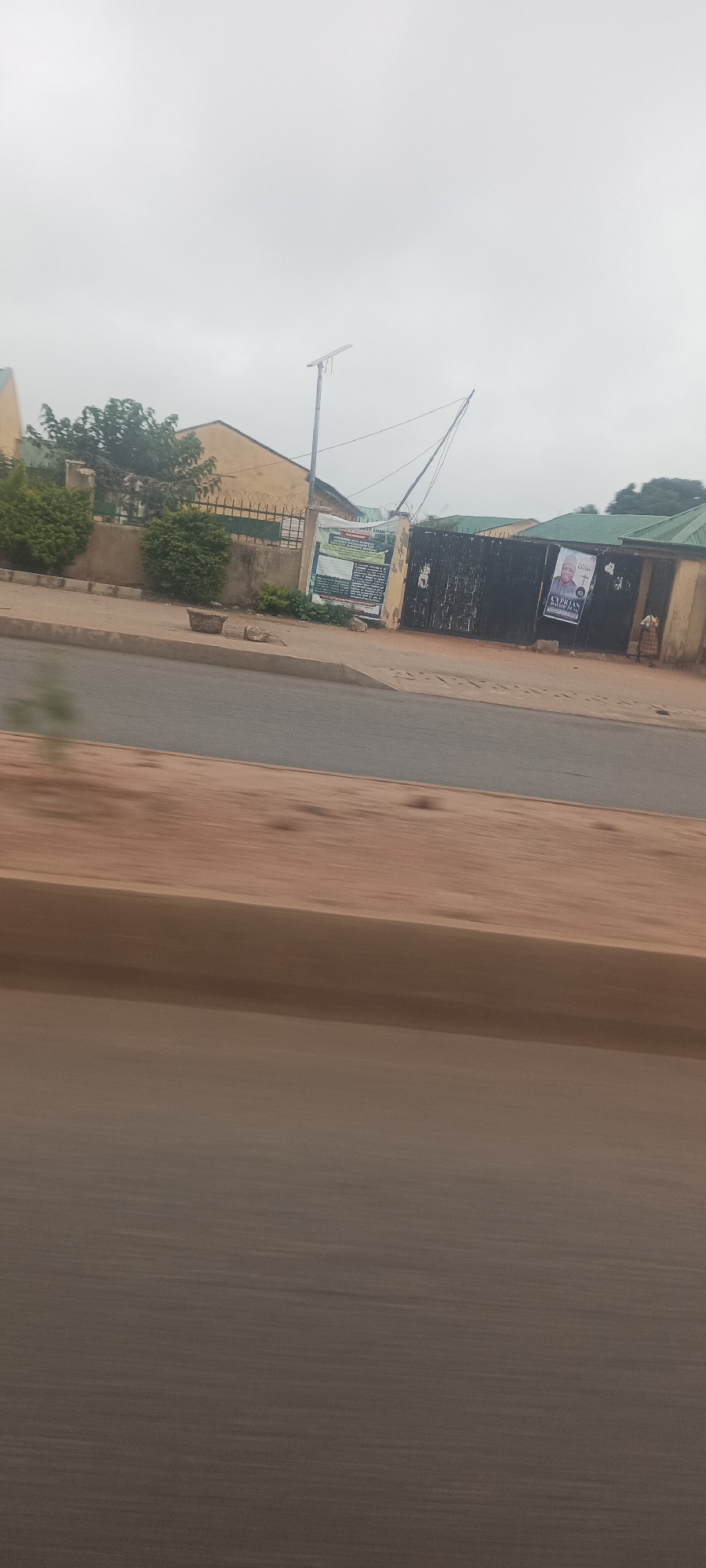 The image presents a blurred roadside view of a street scene in Jos, Nigeria, likely captured from a moving vehicle as suggested by the motion blur across the asphalt road and sandy shoulder in the foreground. Beyond a concrete curb, a perimeter fence topped with barbed wire encloses several single-story buildings with light-colored walls and green roofs, partially hidden by green trees and bushes.
In the midground, a dark metal gate is prominently featured, adorned with two large posters. The poster on the right displays the portrait of a man, with visible text identifying him as "CYPRIAN DANLAMI" and stating "FOR SENATE," indicating a political campaign. A logo is also visible below his name. The poster on the left is a larger, informational banner, predominantly green, white, and black, but its specific text is largely illegible due to distance and resolution.
The sky above is overcast, suggesting a cloudy daytime, and a modern street light with a solar panel is visible against the muted sky, extending above the fence line. No people are visible, and there is no discernible activity taking place beyond the static street and building elements. The overall setting appears to be a semi-urban or residential area.