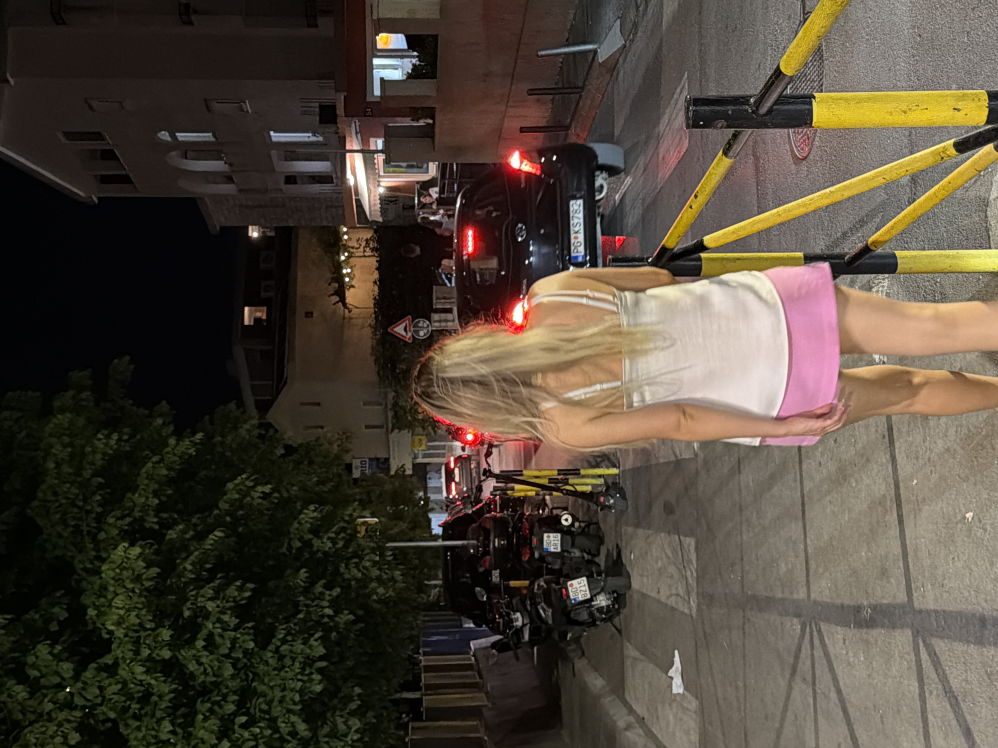 A woman with long blonde hair is walking away from the camera down a street at night. She is wearing a white tank top and a pink skirt. In the background, there are several parked motorcycles and a black car with its brake lights on. The street is lined with buildings and trees. The image appears to have been taken in Budva, Montenegro.
