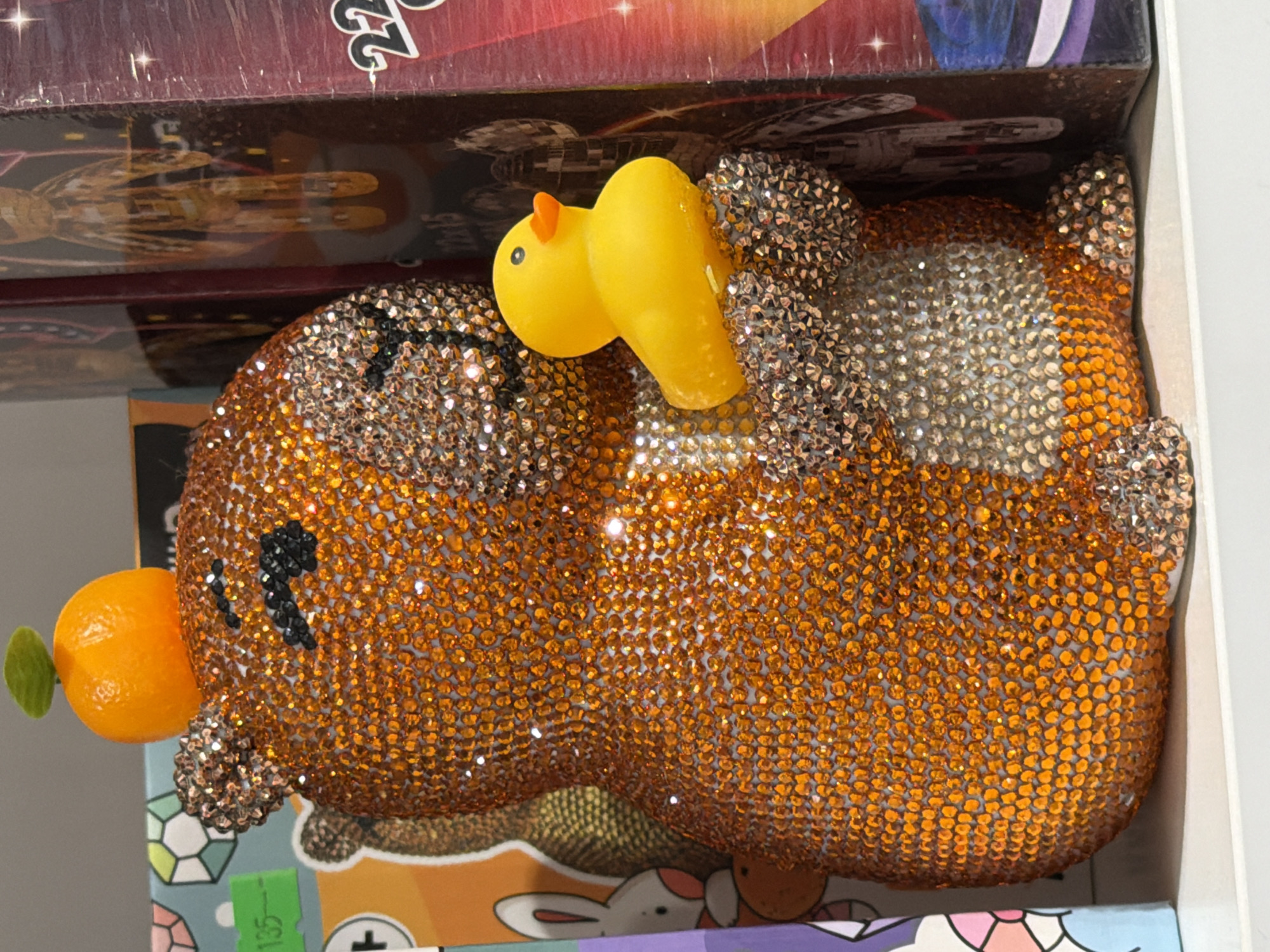 An indoor, eye-level shot captures a vibrant decorative object shaped like a stylized animal, possibly a cat or dog, entirely covered in glittering rhinestones. The animal's body is predominantly adorned with bright orange rhinestones, while its paws and parts of its head feature silver-colored rhinestones. Black rhinestones form its small, stylized eyes and whiskers. Perched on its back is a small, bright yellow rubber duck with an orange beak. On what appears to be its head or snout, there's a small, round, orange fruit-like embellishment with a tiny green leaf, resembling a tangerine.
The decorative animal rests on a colorful surface, possibly packaging, displaying cartoon-like illustrations of bunnies and geometric shapes. A green label with the number "135" is visible on this lower surface. Above the main subject, a dark reddish-brown box is visible, featuring partially obscured text, including the number "22", and an illustration that appears to be a stylized creature or a spaceship-like design, with details suggesting a 'Millennium Falcon' aesthetic. The overall impression is that of items displayed in a retail environment or a personal collection in Izhevsk, Russia, under artificial lighting, as indicated by the sparkle of the rhinestones.