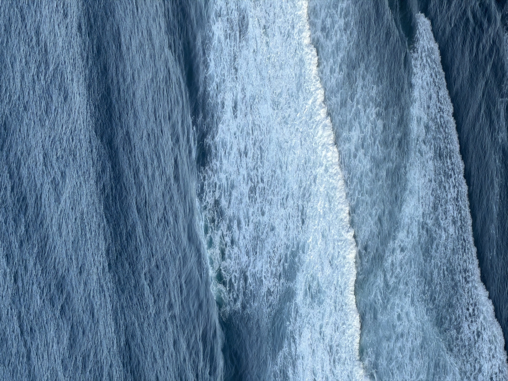 The image shows an aerial view of ocean waves at Kuta Selatan, Indonesia. The waves are breaking, creating foamy white water against the darker blue of the deeper water. The waves appear to be moving from right to left across the frame. The water surface has a textured appearance due to the wave patterns and foam. There are no people or objects visible in the image.