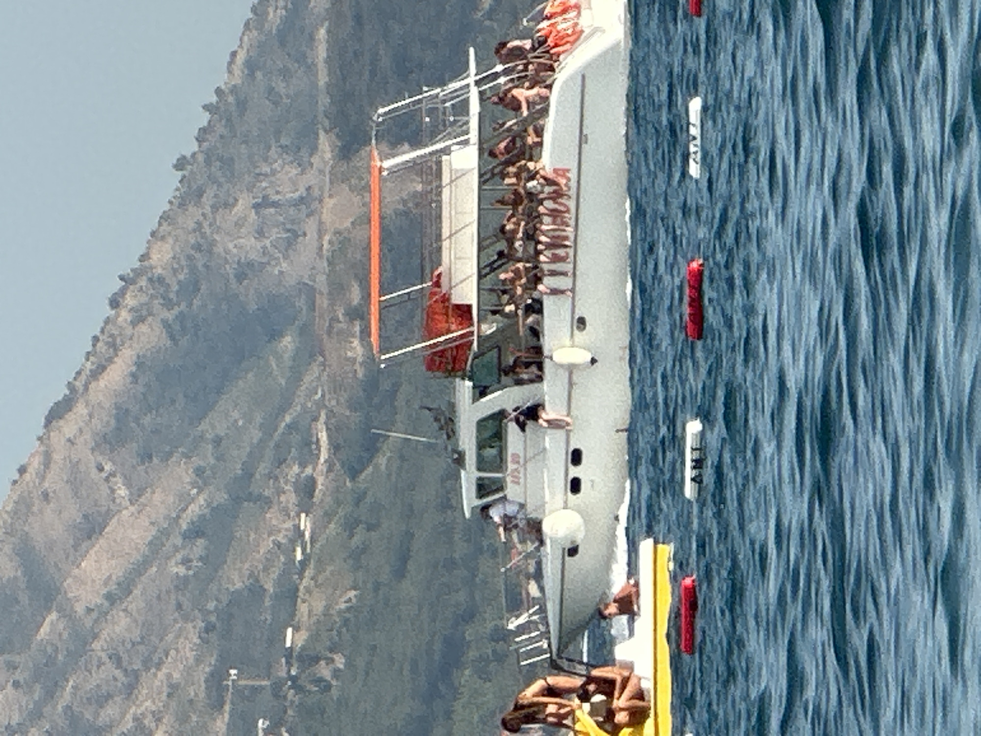 A catamaran-style boat with an orange canopy is sailing on the Adriatic Sea near Boreti, Montenegro. The boat is filled with passengers, some of whom are visible on the deck and in the water. There is a smaller yellow watercraft in the foreground with people on it. The background features a lush, green hillside with some visible structures. Several buoys are floating in the water, indicating a designated swimming or boating area. The water is a deep blue, with visible waves.