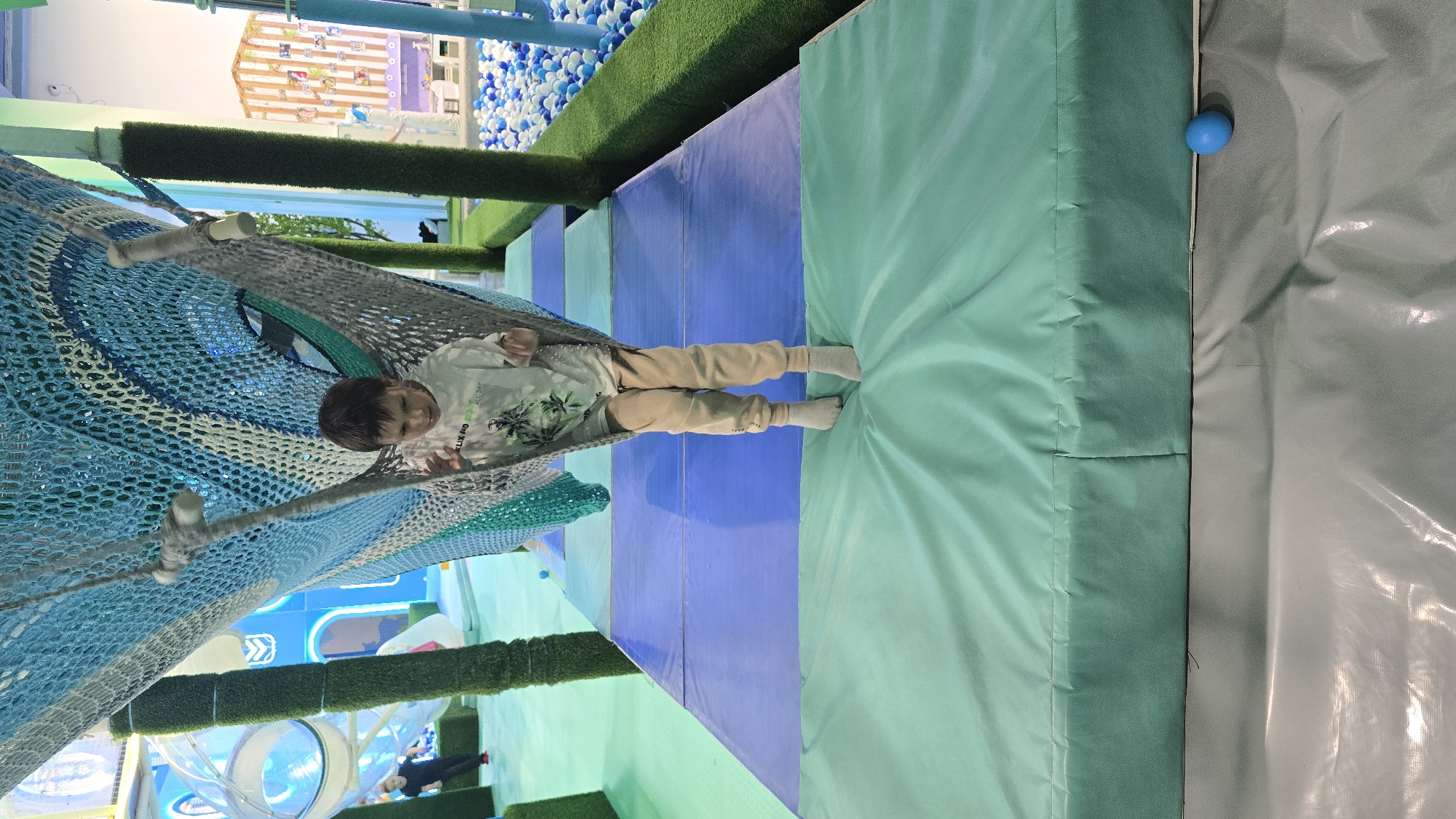 This image depicts a young male child within an indoor children's play area in Izhevsk, Russia. The child, estimated to be between 6 and 10 years old, has short dark hair and is wearing a white, short-sleeved t-shirt with a green palm tree graphic and the text "FELIX ALL DAY" (partially visible). He is also wearing light beige sweatpants and light grey socks.
The child is positioned upright, standing with his feet on a light teal-green soft play mat, while his upper body is enclosed within a suspended, multi-colored net structure. This net structure, primarily composed of blue (various shades), grey, and teal netting, appears to be a tunnel or hammock section of a larger obstacle course. An abstract eye-like design, woven into the blue and teal netting, is visible on the child's right side. The net is supported by grey fabric-wrapped ropes. The child's gaze is directed downwards and slightly to his left.
The floor directly beneath the child consists of a thick, light teal-green soft play mat, which rests on a grey, reflective, plastic-like floor covering. A small, spherical blue ball is situated on this grey covering to the lower right of the child.
Behind the child, the floor extends into a path constructed from alternating rectangular soft play mats in shades of blue and light teal-green. To the immediate right of this path, a border of artificial green turf separates the path from a large ball pit. The ball pit, located in the mid-to-far right background, is densely filled with numerous small blue and white plastic balls.
In the left background, transparent tube slides and tunnels, part of a larger climbing structure, are visible. Several vertical supports, wrapped in artificial green turf, are distributed throughout the play area. A blurred figure of another individual, possibly an adult or older child, dressed in dark clothing, is partially discernible in the far left background near the transparent tubes.
In the far right background, beyond the ball pit, a light-colored, vertically striped structure, resembling a small playhouse facade decorated with various pictures, stands against a white wall. The overall color scheme of the facility emphasizes cool tones of blue, green, and white/grey.