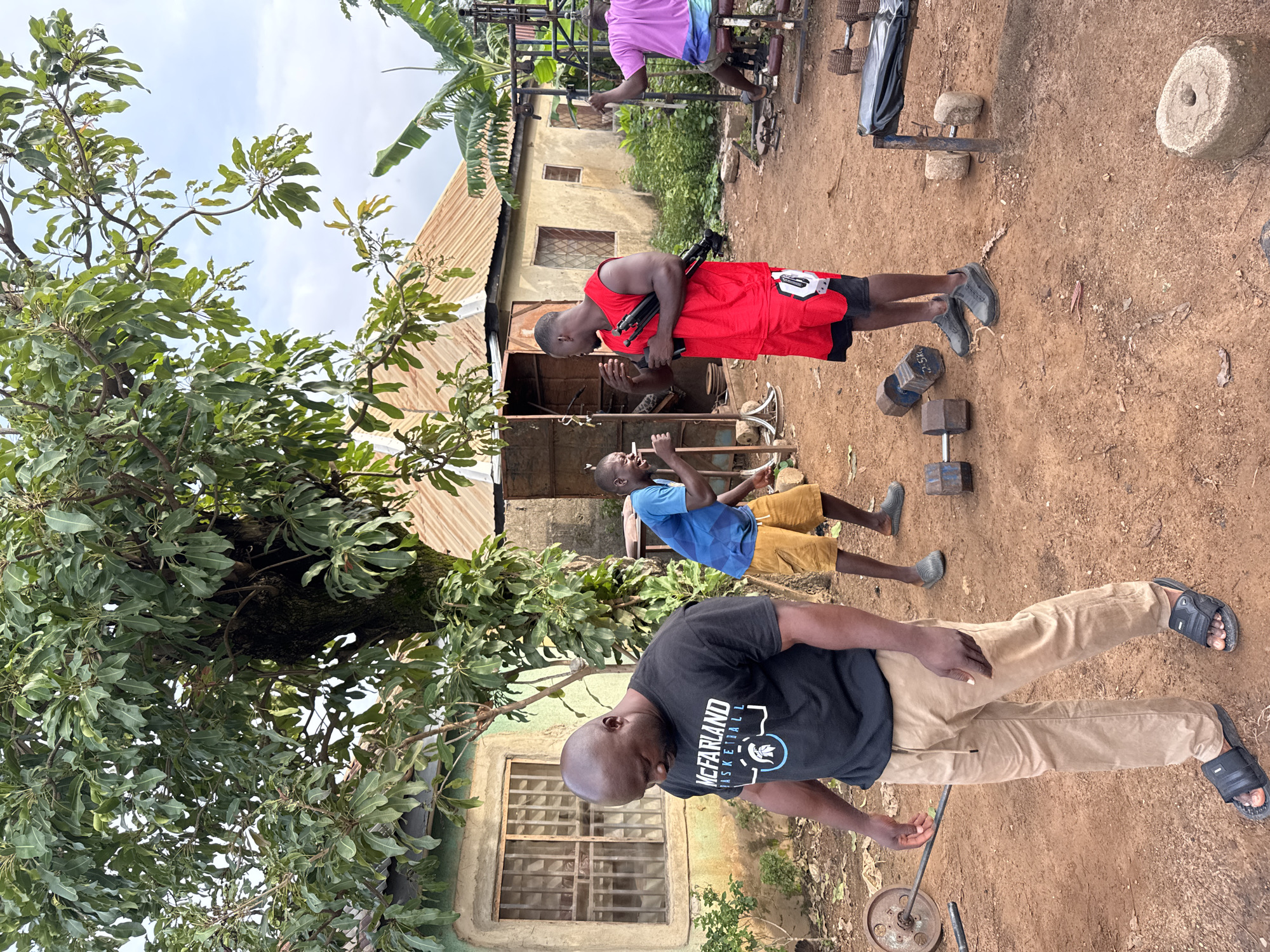 The image depicts an outdoor gym scene in Akwanga, Nigeria. In the foreground, a man wearing a black t-shirt with "MCFARLAND BASKETBALL" and tan pants walks to the left, his head bowed. To his right, a man in a blue t-shirt and yellow shorts is mid-motion, appearing to speak or laugh, with his left arm raised. Further right, another man in a red sleeveless shirt and black and red shorts holds a black object, possibly a tripod or camera stand, with his right arm. Behind him, a man in a pink t-shirt sits on a weight machine. The ground is dirt, with various pieces of exercise equipment scattered around, including dumbbells, a weight bench, and a barbell. Buildings with corrugated metal roofs are visible in the background, along with a large tree providing shade. The environment suggests a casual, community-based fitness setup.