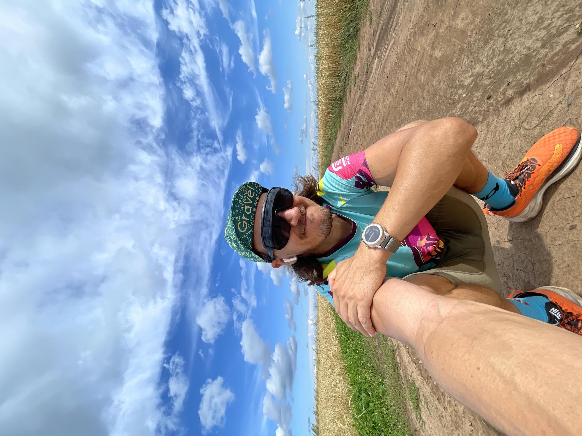 A man is squatting on a dirt path in a field of golden wheat. He is wearing a green baseball cap with white lettering that reads "Gravel," black sunglasses, a teal and pink athletic shirt, khaki shorts, bright orange running shoes, and blue socks with white accents. He has a watch on his left wrist and white wireless earbuds in his ears. His left arm is resting on his right leg. The sky is blue with scattered white clouds. The scene appears to be located in the city of Maksimovo, Russia, based on the presence of a wheat field and dirt path, which are common in rural Russian landscapes.