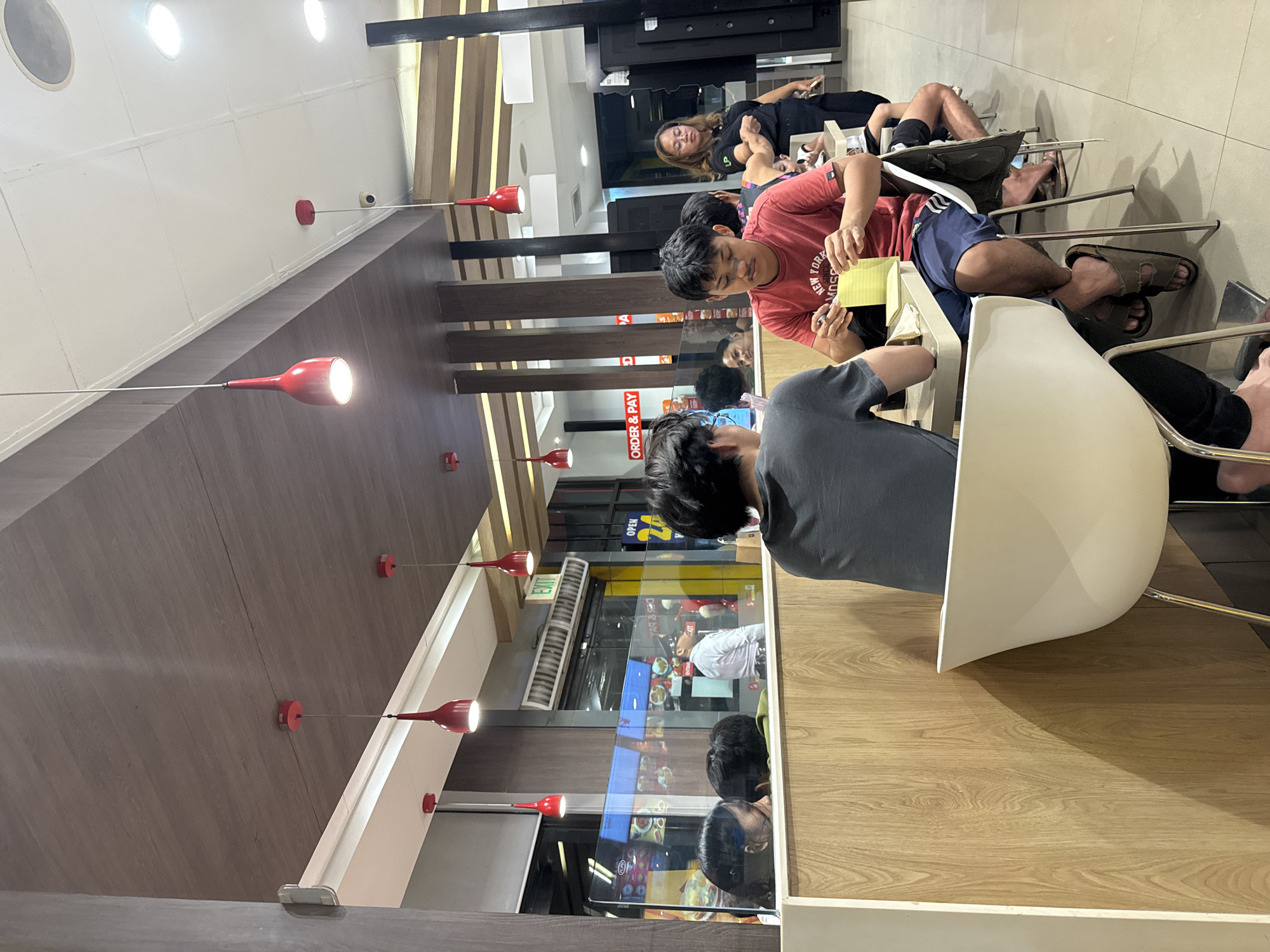 This media file captures an indoor scene within a casual dining establishment, likely a fast-food restaurant or eatery, in Davao City, Philippines. The perspective is slightly elevated and angled, looking across a row of tables and seated patrons.
In the foreground, a person in a dark grey t-shirt is partially visible from behind, seated at a light wooden table. Next to them, a young man in a red t-shirt, which has "NEW YORK SUMMER" printed on it, is actively writing or sketching on a yellow notepad. Other individuals are seated further back; a woman with long dark hair wearing a black top is visible in the mid-ground, and several pairs of legs and feet, including those wearing sandals, are seen beneath the tables.
The decor is modern and clean, featuring light wood-paneled walls and table surfaces, contrasted with dark wooden beams and accents. Red, conical pendant lights hang from the ceiling, providing bright illumination, suggesting it could be evening or an enclosed space. A glass partition separates the dining area from a service counter or kitchen, where a prominent red sign clearly reads "ORDER & PAY" in white text. An "EXIT" sign is also visible on a wall, and a reflection in the glass shows a partially visible "OPEN" sign and a person in a white shirt. A small security camera is mounted on the ceiling. The overall activity suggests people are either waiting for their orders, dining, or casually passing time.