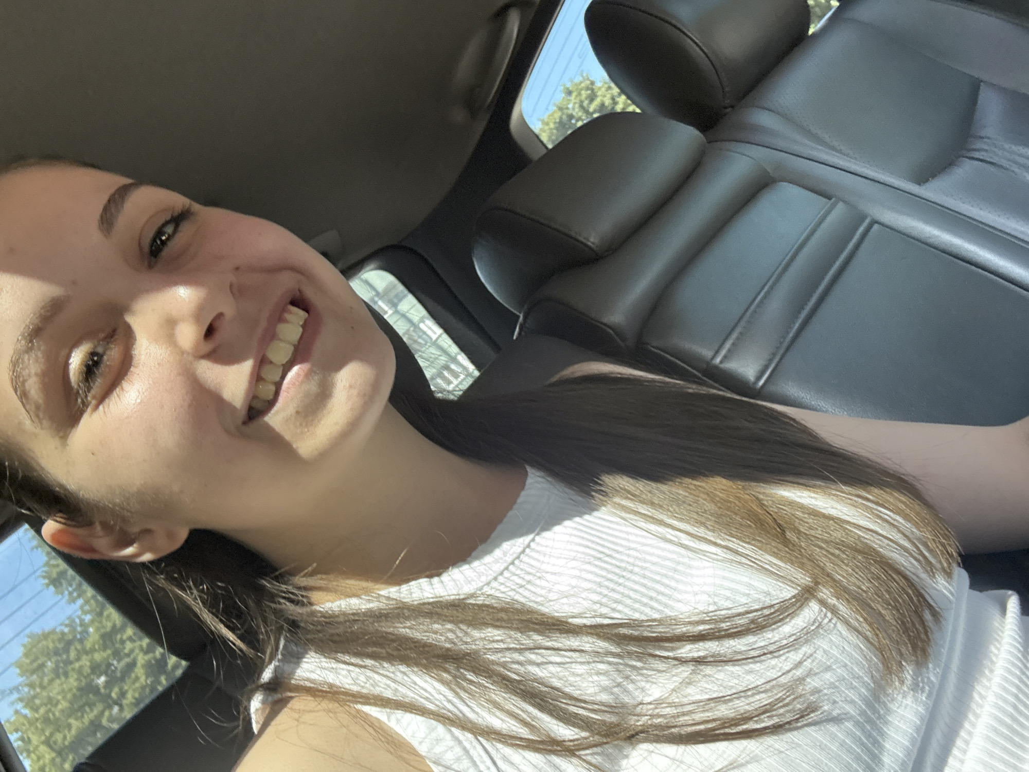 The image is a brightly lit selfie taken by a young woman inside a car, likely in Burgas, Bulgaria. She is seen from the chest up, smiling broadly with her teeth showing, and her eyes are open. Her long, dark brown hair falls over her shoulders and a white, ribbed top. Strong sunlight illuminates her face from the right, casting shadows on her left cheek and forehead, suggesting direct sun exposure.
The setting is the interior of a car, with dark-colored, possibly leather, seats visible behind her and to her right. Through the car windows, a clear blue sky and abundant green foliage from trees are visible, indicating sunny, clear weather during daytime. The overall mood conveyed by her expression is one of happiness or cheerfulness. No text is visible in the photograph.