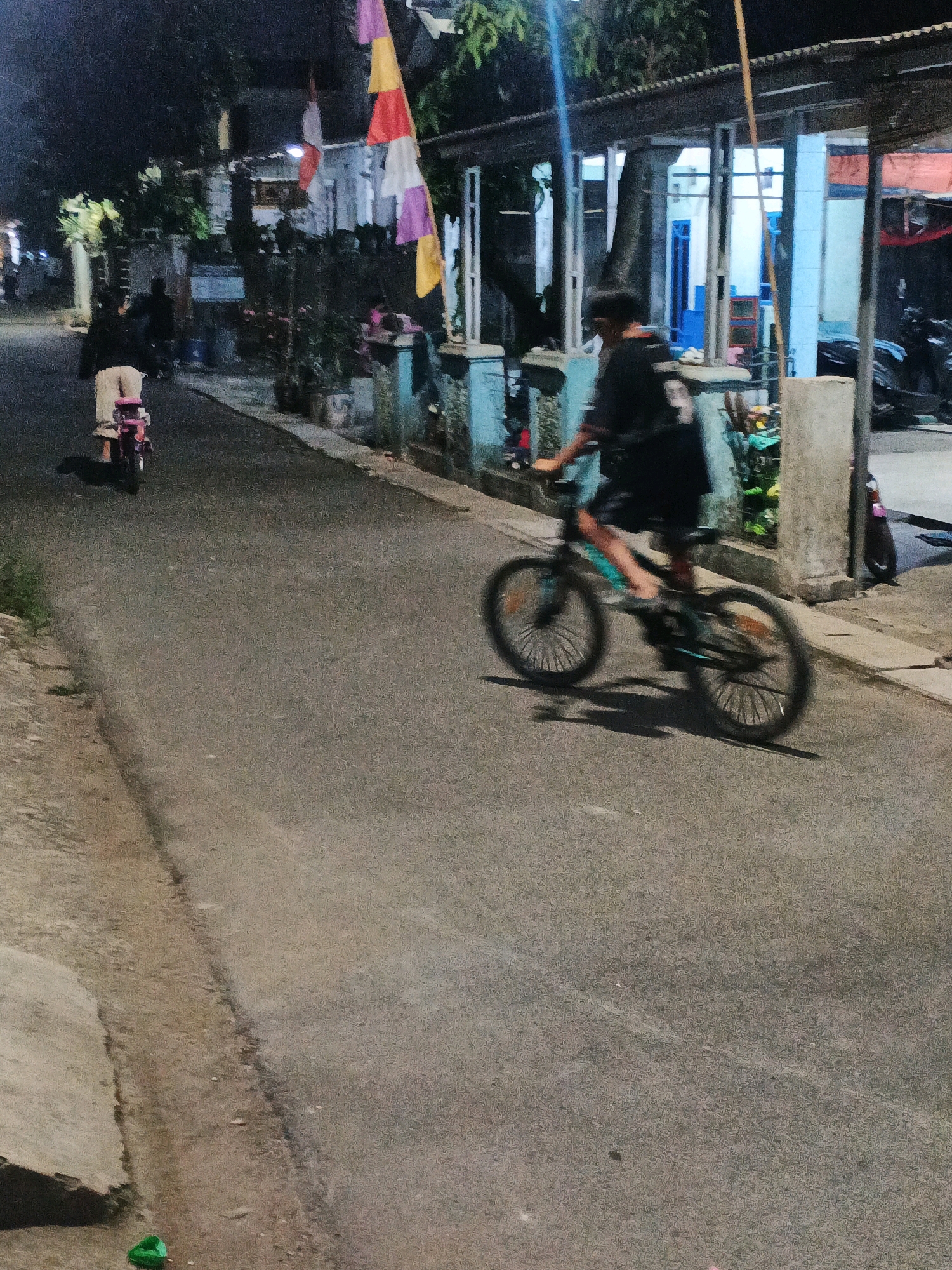 The image shows a street scene at night in Kadu, Indonesia. A person is riding a bicycle in the foreground, their motion blurred by a long exposure. The cyclist is wearing a black shirt and shorts. To the left, another person is riding a pink child's bicycle. In the background, there are houses and trees. Colorful flags are hanging from a line across the street. The street is paved and appears to be a residential area.