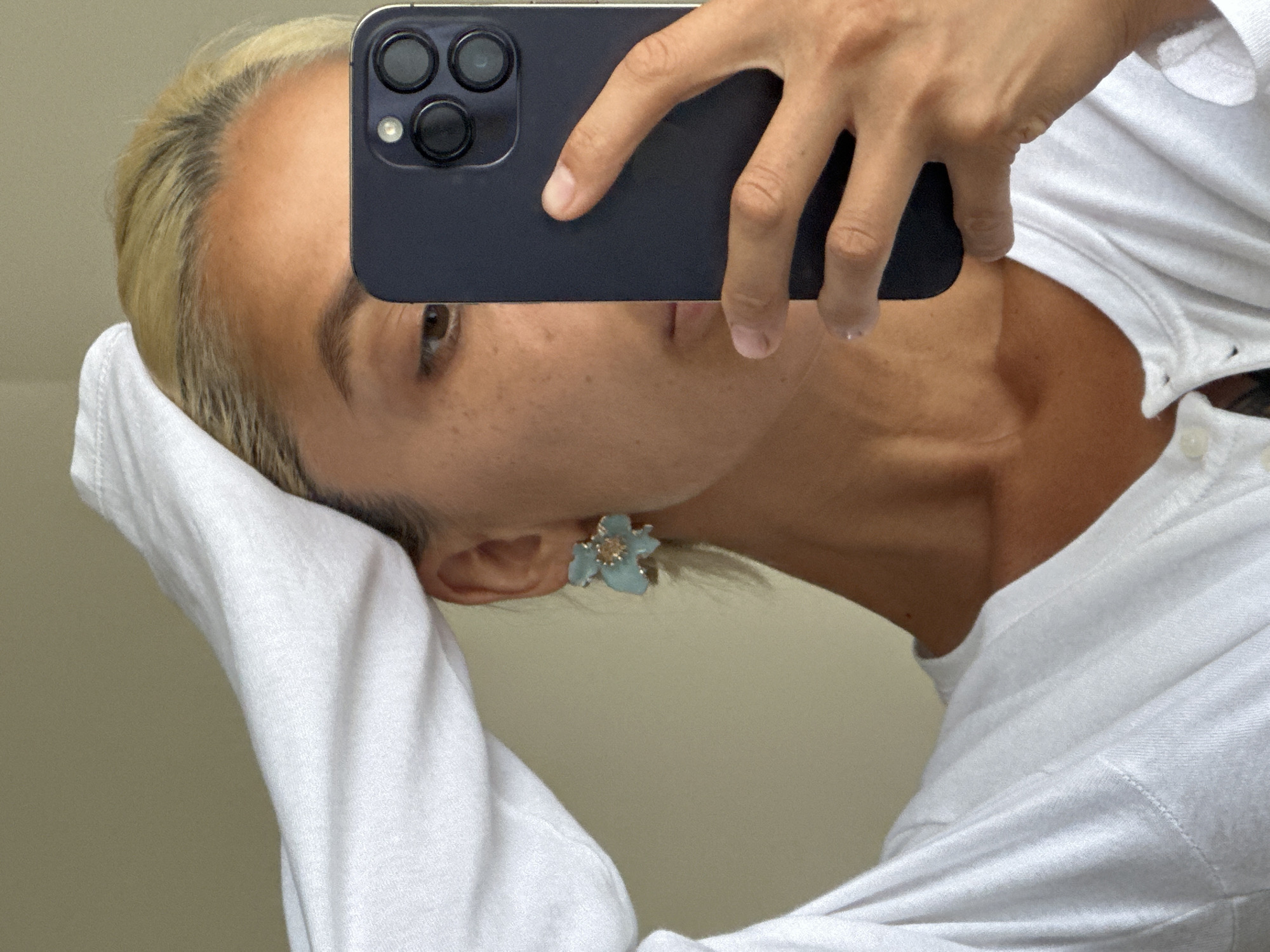 A person is taking a selfie in what appears to be a hotel room in Kuta Selatan, Indonesia. The person has blonde hair and is wearing a white long-sleeved shirt. They are also wearing a light blue flower-shaped earring. The person is holding up a smartphone, which is obscuring part of their face, and their left hand is resting on the back of their head. The background is a plain, light-colored wall.