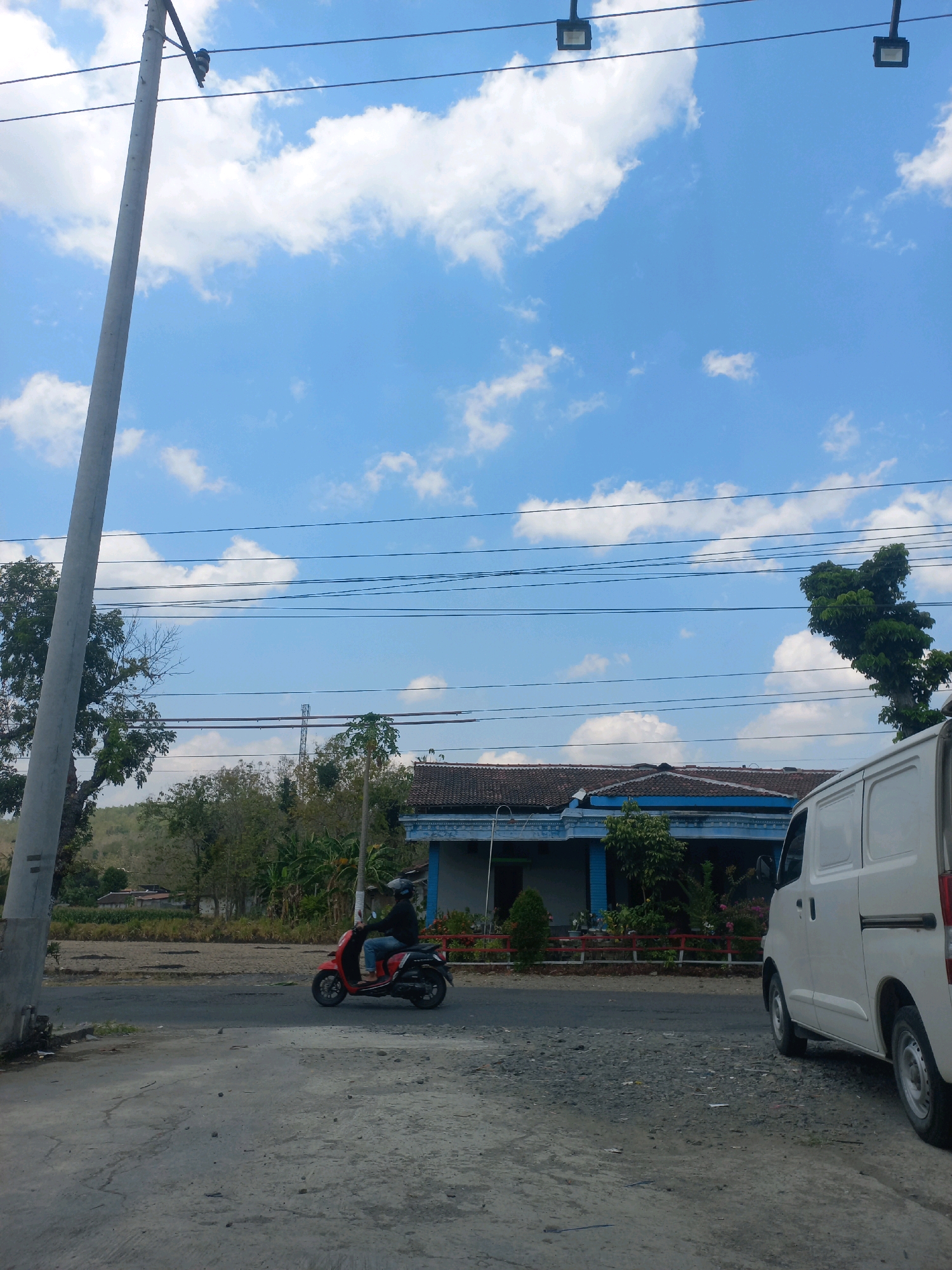 A person rides a red scooter along a road in Baturetno, Indonesia. The person is wearing a black jacket and blue jeans. A white van is parked on the right side of the road. In the background, there is a house with a blue roof and a red fence. The sky is blue with white clouds. The image appears to have been taken on a sunny day.