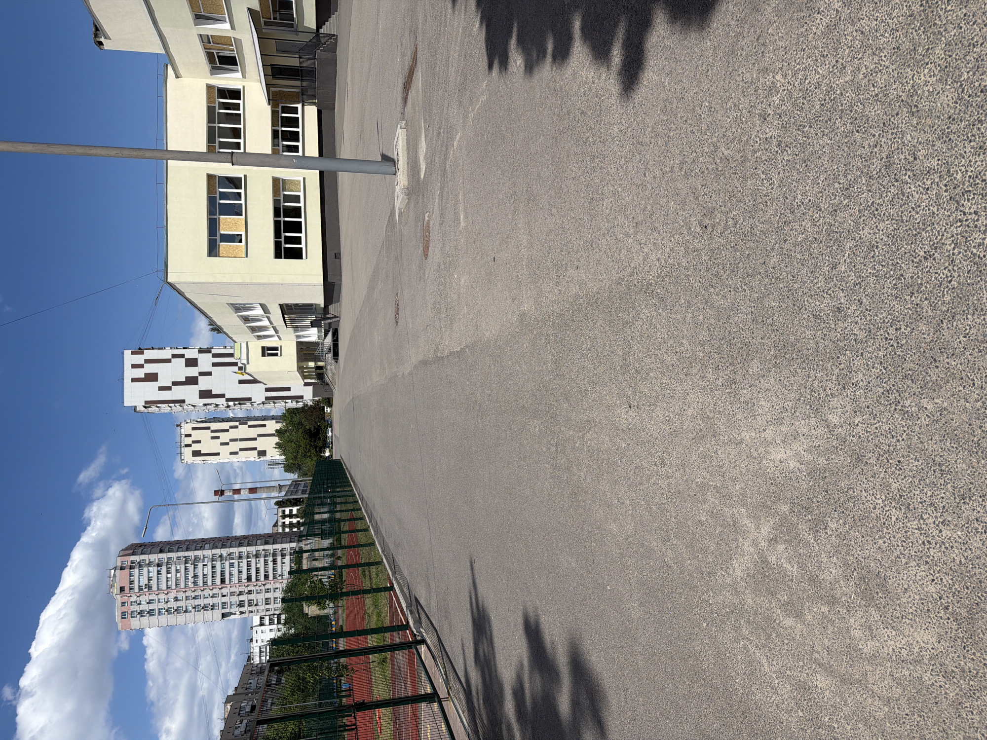 The image depicts an outdoor scene with a focus on a paved area in the foreground and buildings in the background. The paving appears to be asphalt, with some visible texture and slight discoloration. Shadows of people and trees are cast across the pavement, suggesting it is daytime. A metal pole stands on the left side of the paved area, with some wires connected to it.
In the background, several buildings are visible. The closest building is light yellow and features multiple windows. Further back, there are modern residential buildings with distinctive architectural patterns and colors. One building is tall and slender, with many windows. Another is a complex of interconnected structures with a checkered facade. A green fence is visible along the edge of the paved area, separating it from a sports field with a red track.
The sky is blue with scattered white clouds, indicating fair weather. The overall scene suggests an urban environment, likely a schoolyard or a public space adjacent to residential and educational facilities. The orientation of the image is sideways. No text is visible in the image.