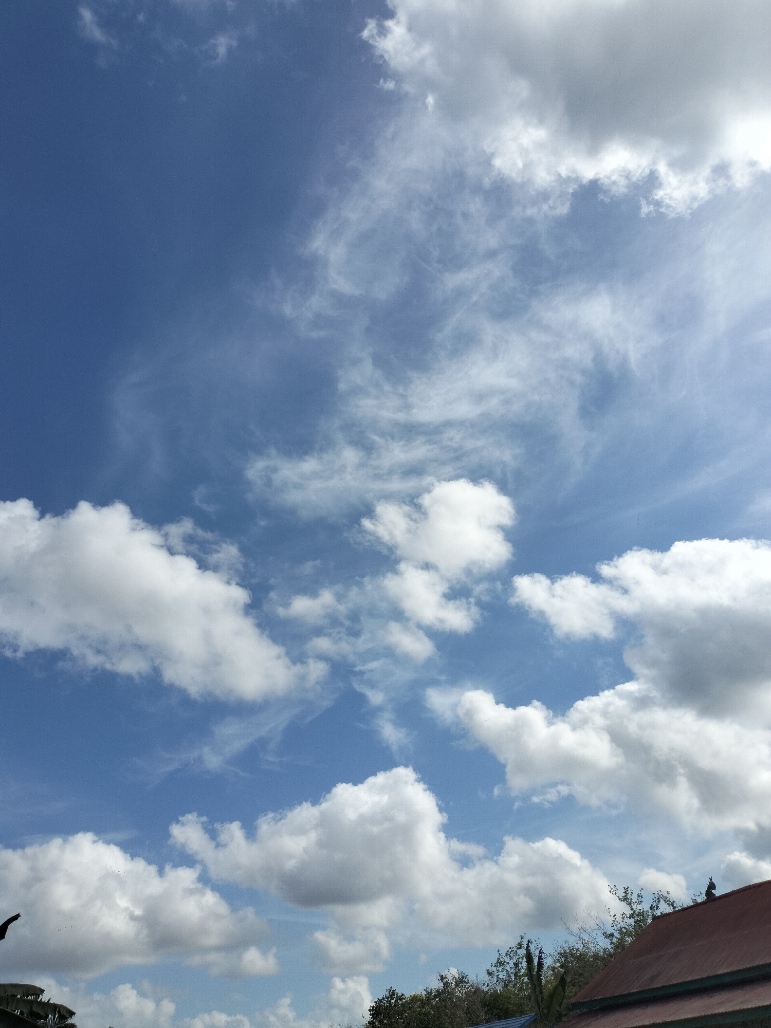 This is a vertical, eye-level, outdoor, daytime shot of a blue sky with scattered clouds and a portion of a rooftop in the foreground. The sky is mostly clear with wispy cirrus clouds in the upper half and larger, cumulus clouds lower down. A small section of a reddish-brown tiled roof is visible on the right side of the frame, with trees and foliage just below it. Two dark silhouettes, possibly birds or small figures, perch on the roof. A hint of a blue structure is visible on the far left, partially obscured by foliage. The image appears to be taken in Karang Indah, Indonesia, likely from a slightly elevated position.