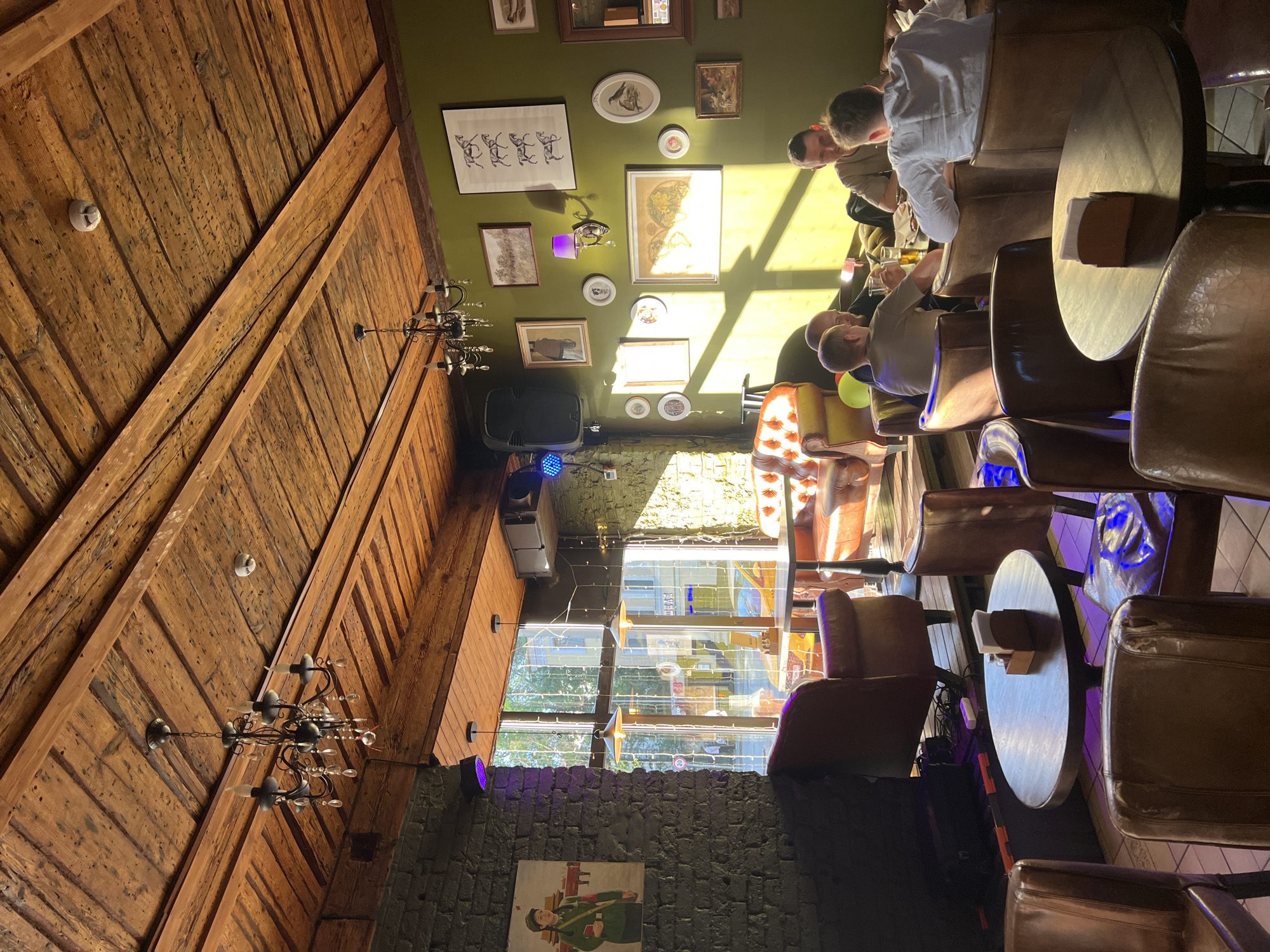 This image displays the interior of a commercial establishment, specifically a bar or cafe, located in Moscow, Russia, as evidenced by the mirrored Russian word "МАГАЗИН" (SHOP) visible on a building through the large window. The space is characterized by a rustic aesthetic, featuring a ceiling constructed from dark, distressed wooden planks and exposed timber beams. Two multi-arm metal chandeliers, adorned with faux candles and crystal drops, hang from the ceiling, alongside a white smoke detector.
The left wall is composed of dark gray-painted brick and displays a framed poster depicting a woman in a green military-style uniform with a red arm band. A large black speaker is mounted high on this wall. The right wall is painted a dark olive-green and is densely arrayed with various framed artworks, including depictions of horses, birds, maps, and landscapes. A wall-mounted sconce with a purple lampshade is also present on this wall.
Natural sunlight streams through the large window, casting bright highlights and distinct shadows across the floor and the green wall. Artificial lighting, including spotlights (one emitting purple, another blue light) and the chandeliers, supplement the ambient illumination.
The furniture primarily consists of dark brown leather armchairs and chairs, some exhibiting wear, arranged around dark wooden tables of various shapes. In the right midground, four adult males are seated around a rectangular table, engaged in conversation. One individual, wearing a light-colored shirt, is positioned with his back to the camera, while another, wearing a t-shirt, holds a glass. A yellow leather Chesterfield-style sofa is partially visible in the left midground, near the window. A small wooden napkin dispenser rests on a round table in the foreground.