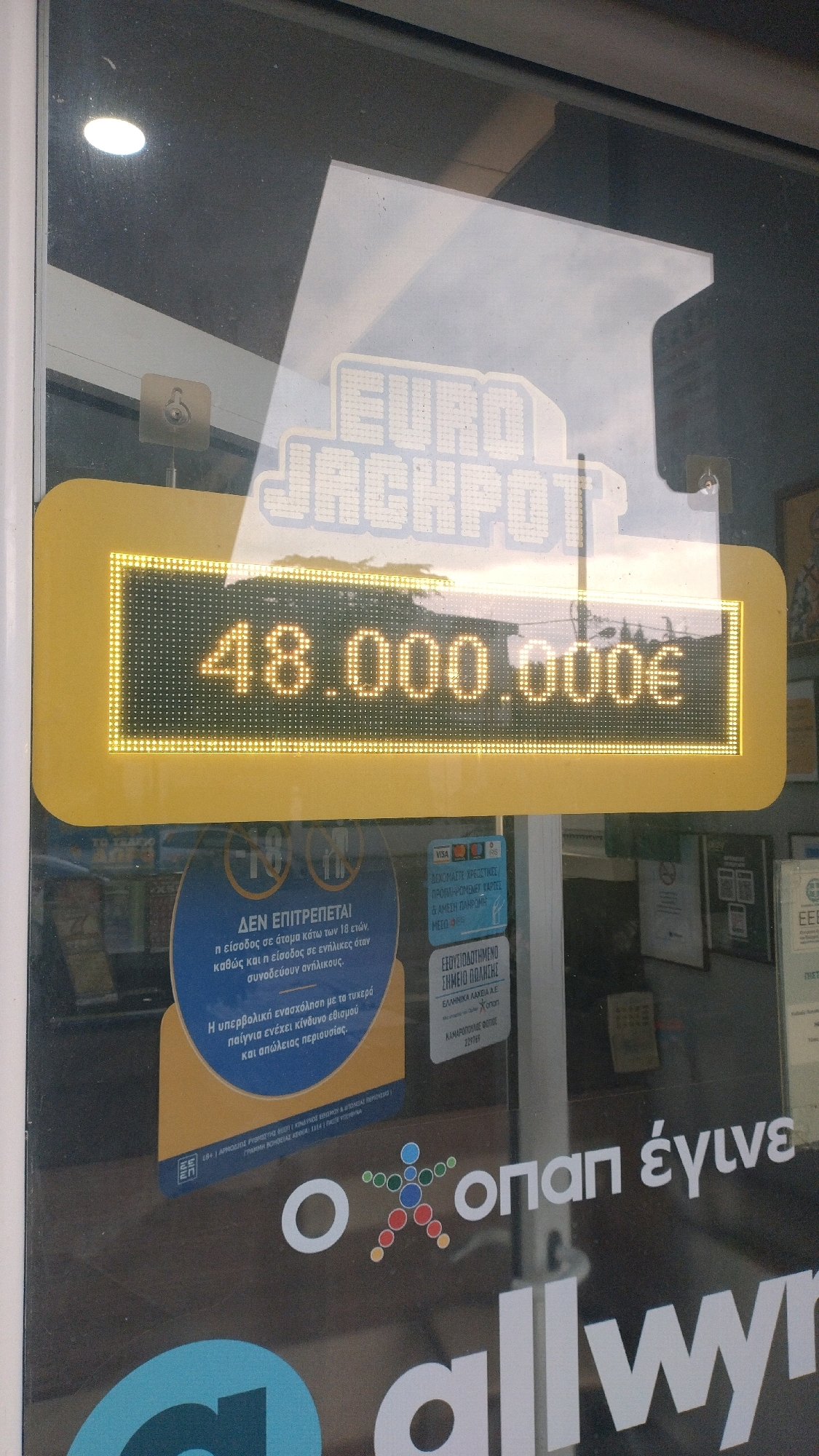 The image shows a yellow illuminated sign with "EURO JACKPOT" in large white letters above a digital display showing "48.000.000€". The sign appears to be in the window of a shop. Below the sign, there is a blue circular sticker with Greek text and a warning symbol indicating it is not for persons under 18. Further down, there are other stickers and some text on the glass, including logos for VISA and Mastercard, and Greek text that translates to "gambling can be addictive". The text "ΟΠΑΠ έγινε" (Opap became) is visible on the window below the stickers, with a colorful graphic of stylized figures. The reflection in the window suggests an outdoor environment with trees and sky. The specific location is identified as Neoi Epivates, Greece, based on the presence of a sign that reads "EEOYYCIODOTHMENO SHMEIO PAIXTHE" which translates to "Authorized Betting Point". The image was taken from outside the shop, looking in. There are no people visible in the image.