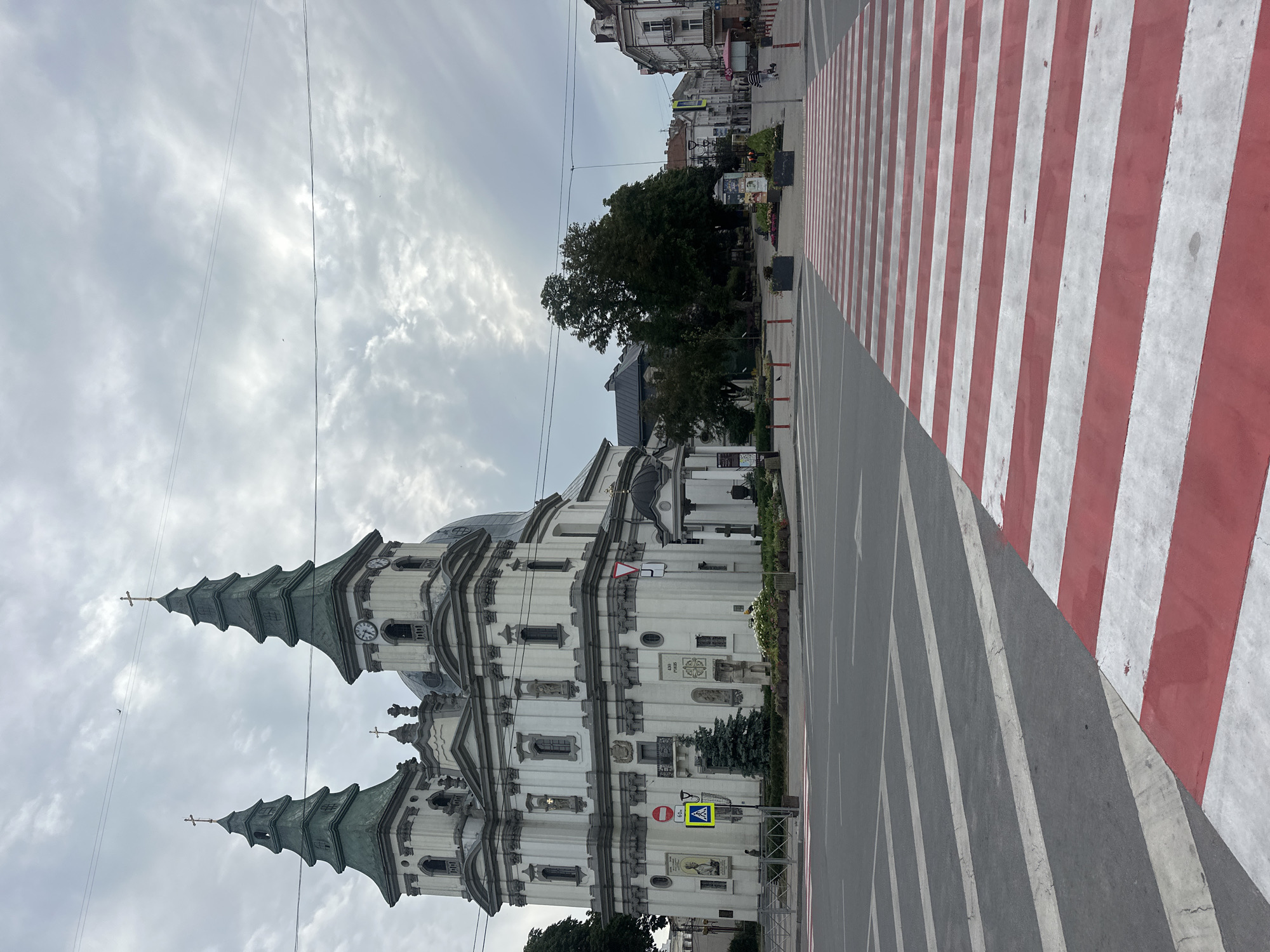 The image shows a view of the Ternopil Cathedral (Nativity of Christ Cathedral) in Ternopil, Ukraine. In the foreground, a pedestrian crossing with red and white stripes is visible on an asphalt road. The cathedral, an ornate white building with green spires and crosses, dominates the left side of the image. The sky is overcast with scattered clouds. Trees and other buildings are visible in the background, suggesting an urban setting. The scene appears to be a public square or intersection in front of the cathedral.