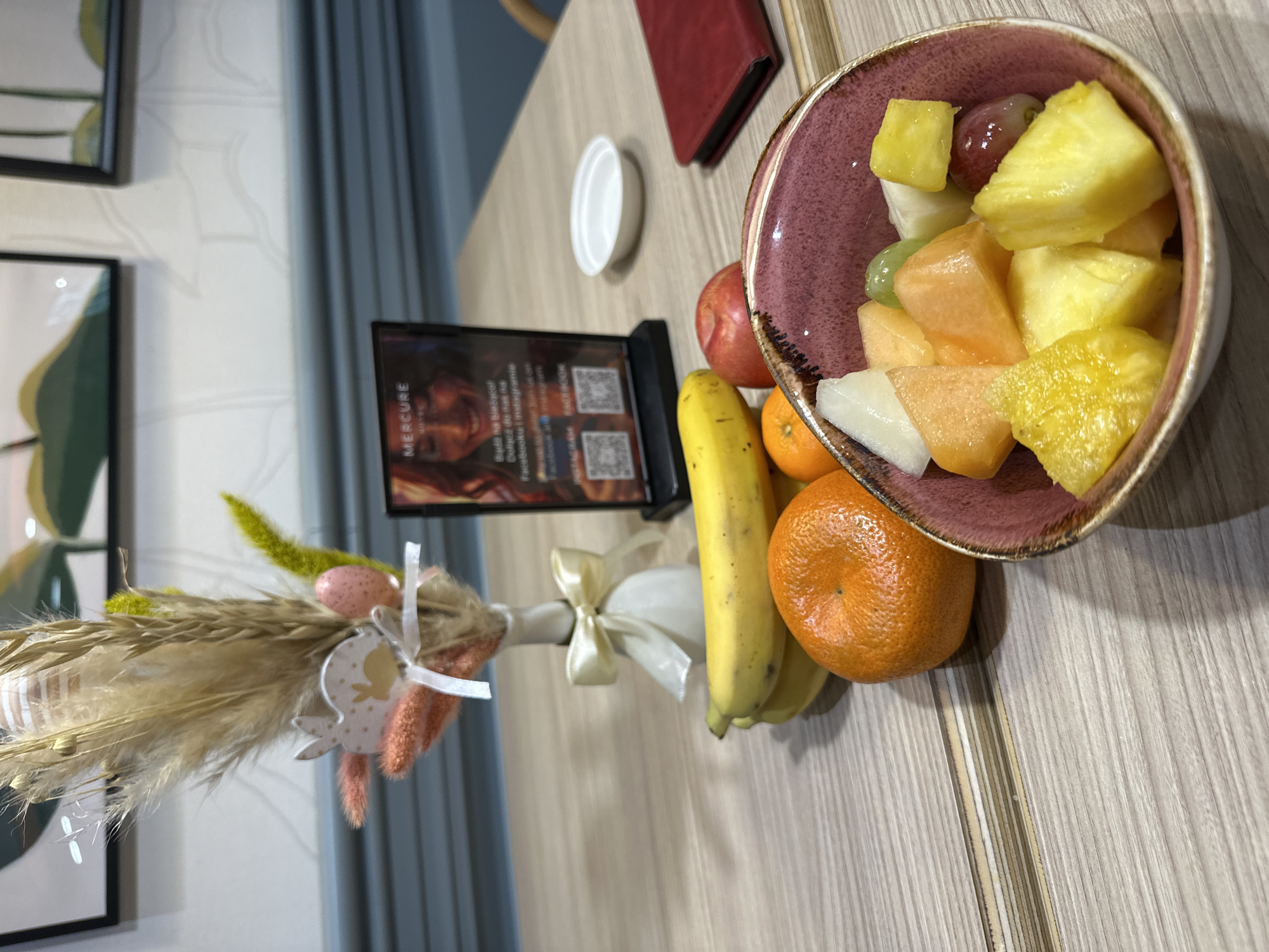 The image shows a table setting in what appears to be a hotel or restaurant, likely in Wrocław, Poland, given the "Mercure" branding visible on a small promotional display. The table has a light wood-grain surface.
The foreground objects include a bowl filled with cut fruits (pineapple, melon, grapes), two tangerines, a banana, a small red object (possibly a menu or book), and a small white dish. Next to the promotional display is a decorative Easter-themed bundle of dried grasses and artificial eggs, tied with a ribbon.
In the background, partially visible, are framed pictures and a blue curtain or wall panel. The scene is well-lit, suggesting indoor lighting. There is no evidence of other people or significant action depicted in the image.