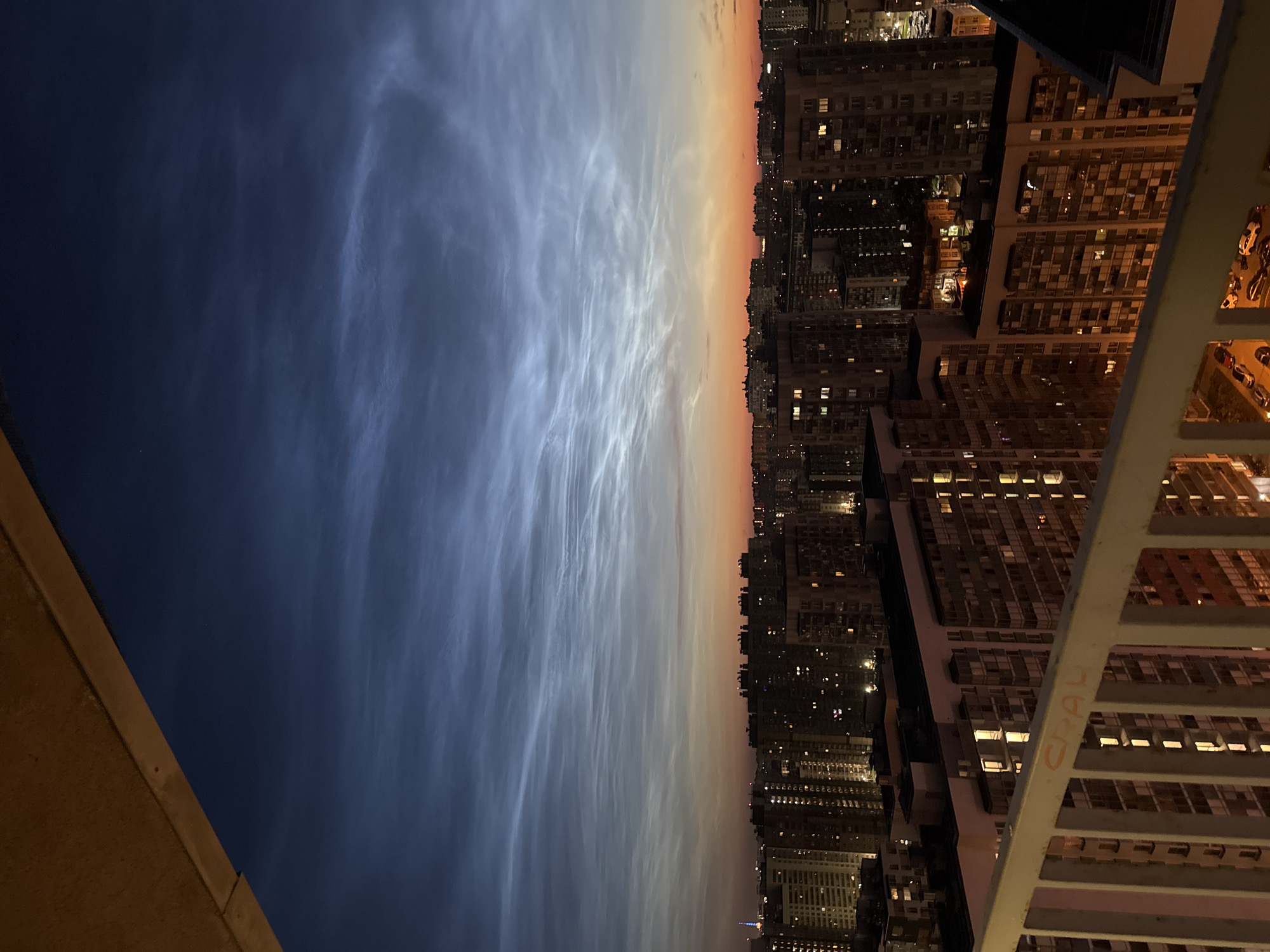 The image depicts a city skyline at dusk, likely in Oreshek-3, Russia, given the context. The sky is dominated by noctilucent clouds, which appear as wispy, luminous formations against a gradient of deep blue and warm orange hues. Below the sky, a dense arrangement of multi-story apartment buildings fills the midground, their windows illuminated with warm, yellow light, indicating occupancy. The foreground features a portion of a balcony railing, painted white and showing some signs of graffiti, with the letters "CRAL" visible in orange. Cars are parked in an outdoor lot visible in the lower right of the image. The overall impression is one of urban life unfolding under an extraordinary atmospheric phenomenon.
