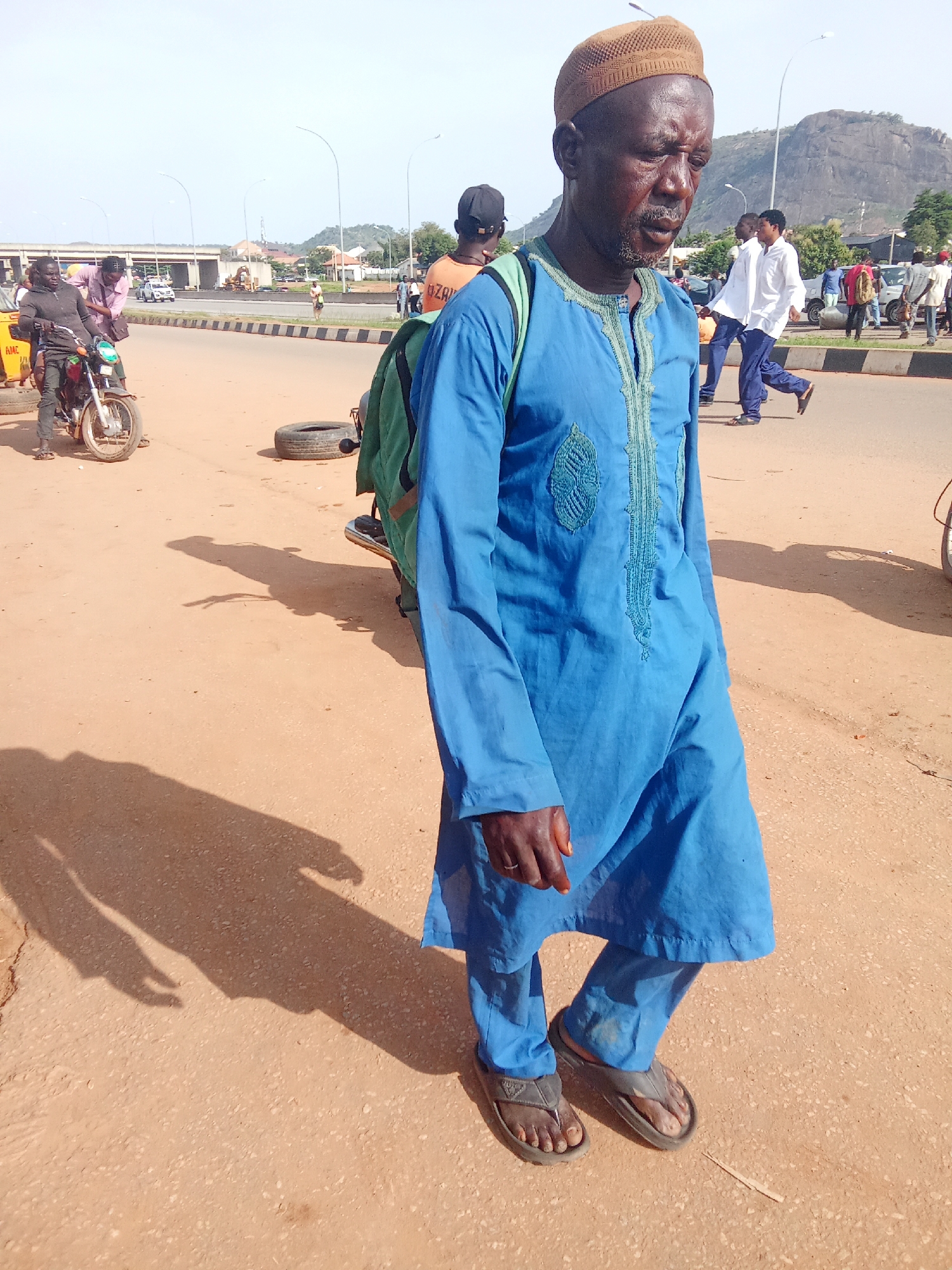 A middle-aged man wearing a brown cap, blue robe, and blue pants walks on a dirt road. He has a green backpack on his left shoulder. The man is looking down and to his right. Behind him, several other people are walking on the road, and a motorcycle is parked on the left. In the background, there is a highway overpass, some buildings, and a rocky hill. The sky is clear and bright.