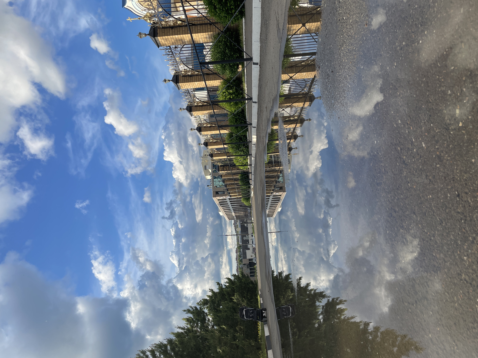 The image shows a reflection in a puddle on an asphalt surface. The reflection captures a blue sky with wispy clouds and a building facade with a fence. The location is identifiable as Izhevsk, Russia, based on architectural and urban features. A tree is visible at the bottom edge of the reflection. The puddle's surface reflects the scene, creating a mirrored effect. The asphalt surface is dark and granular, contrasting with the reflected sky and building.