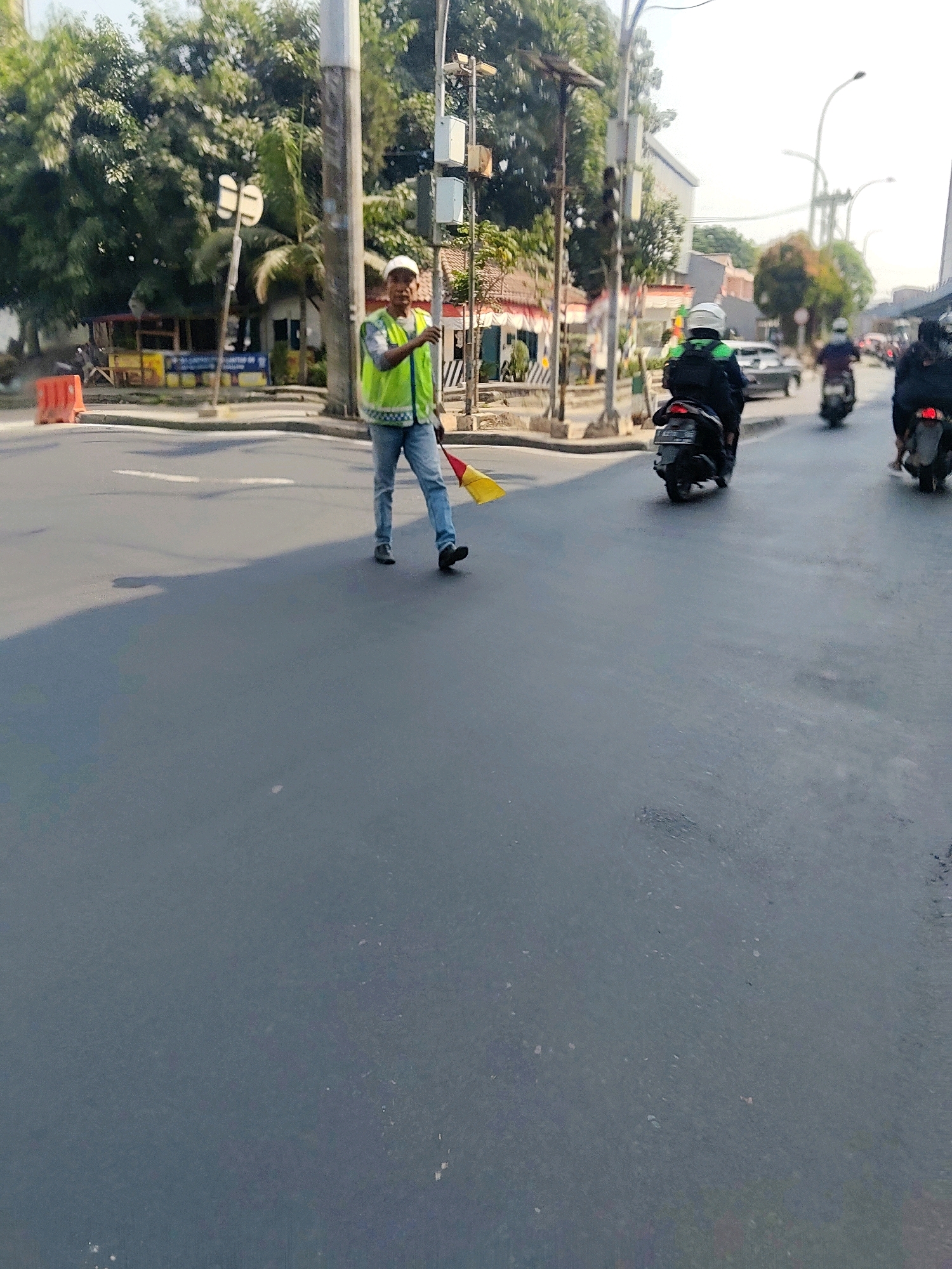 A person in a reflective vest and jeans stands on a paved road, holding a yellow and red flag stick. The person is wearing a white cap. Behind the person, several motorbikes are moving on the road, some with riders wearing helmets. The environment appears to be an urban street with buildings and trees visible in the background. Orange traffic cones are present on the side of the road. The scene is likely an intersection or a street in Pasirsari, Indonesia, given the context provided, where traffic management or assistance is being provided.