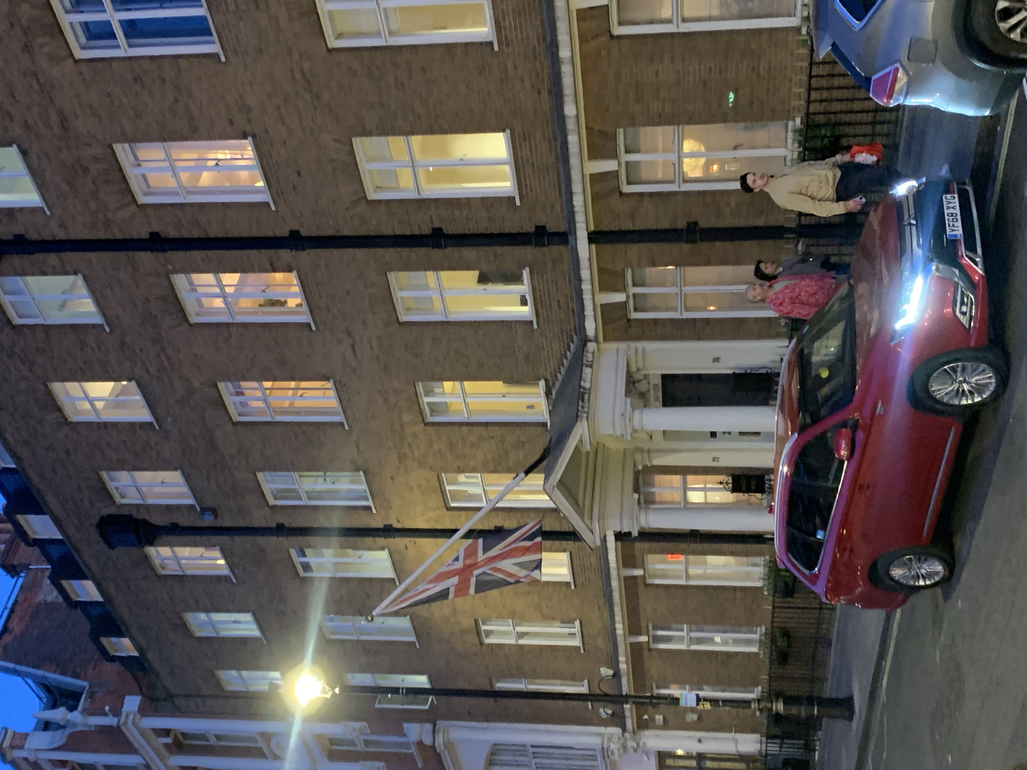 The image captures an evening street scene, rotated so buildings appear horizontal. A multi-story brick building with numerous illuminated windows dominates the background. A Union Jack flag, indicating a British location, hangs from a pole attached to this building. Adjacent to it, another building features an entrance with white columns.
In the foreground, a bright red SUV or car is parked on the street, its headlights shining brightly. The license plate "YF68 XYG" is clearly visible on the red car. Part of a silver car is also visible further down the street.
Several people are gathered near the red car. A young man, dressed in a light-colored jacket and dark trousers, stands upright on the pavement next to the vehicle, looking towards the viewer. He appears to be holding a small object in his left hand. Two other individuals are partially obscured by the front of the red car; one wears a noticeable red patterned top. The group appears to be standing or waiting on the street.
The scene is set at dusk or night, evidenced by the dark sky, the glowing streetlamp on the left, and the interior lights emanating from the building windows, as well as the illuminated car headlights. The overall atmosphere is one of a quiet street scene under artificial light.