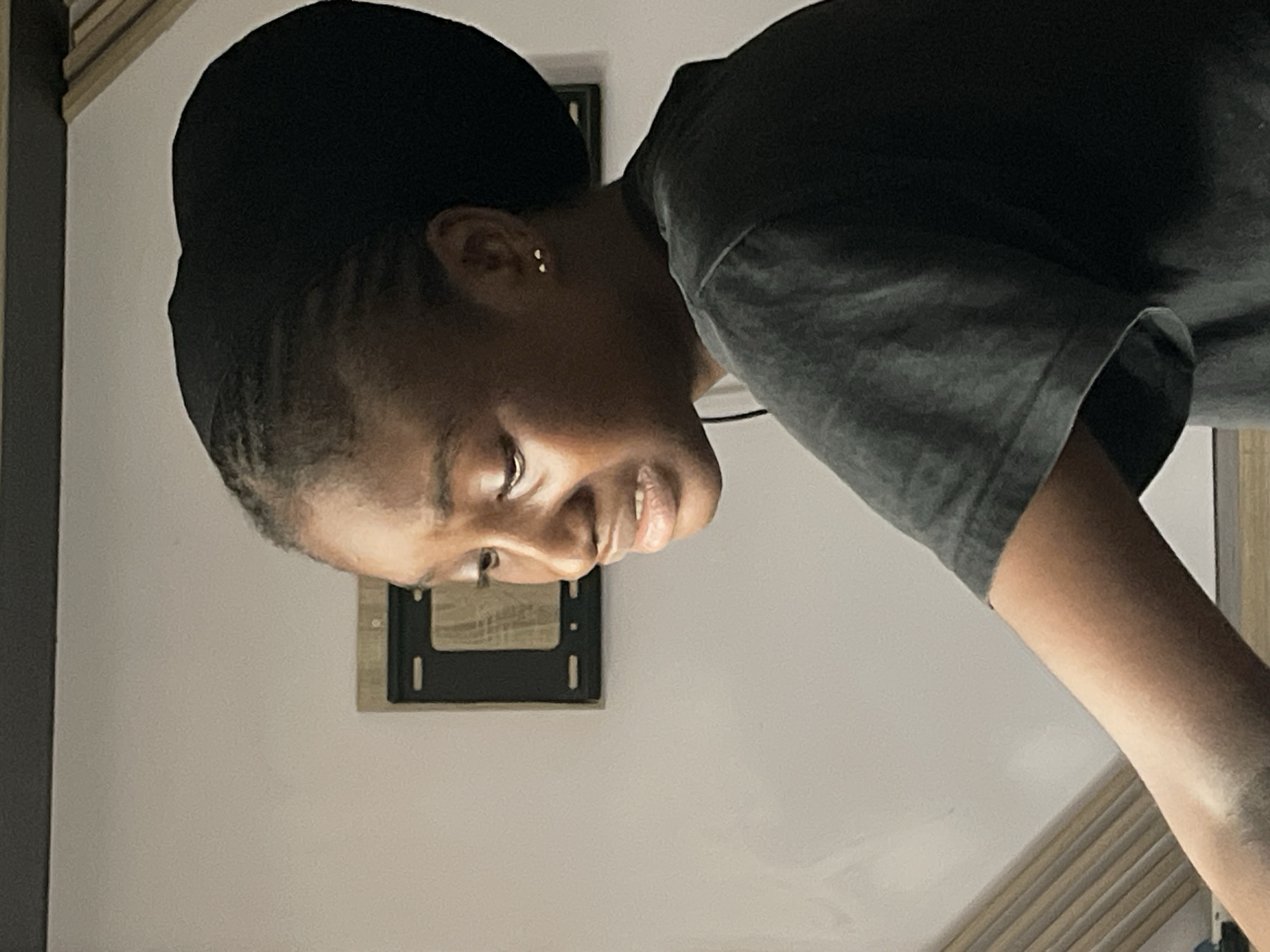 A close-up, medium shot captures a young Black woman from the chest up, facing left and looking downwards. She is wearing a dark grey t-shirt and a black headscarf that covers her braided hair. The woman has her mouth slightly open and her eyes are downcast, giving the impression she might be crying or experiencing sadness. She has a small gold stud earring in her visible right ear. In the background, a light-colored wall is visible, with a dark rectangular object mounted on it, possibly a framed picture or a small screen. To the left of the woman, there are vertical wooden slats. The lighting suggests an indoor setting, possibly a room with ambient light. The image does not provide specific details to identify a location within Abuja, Nigeria.
