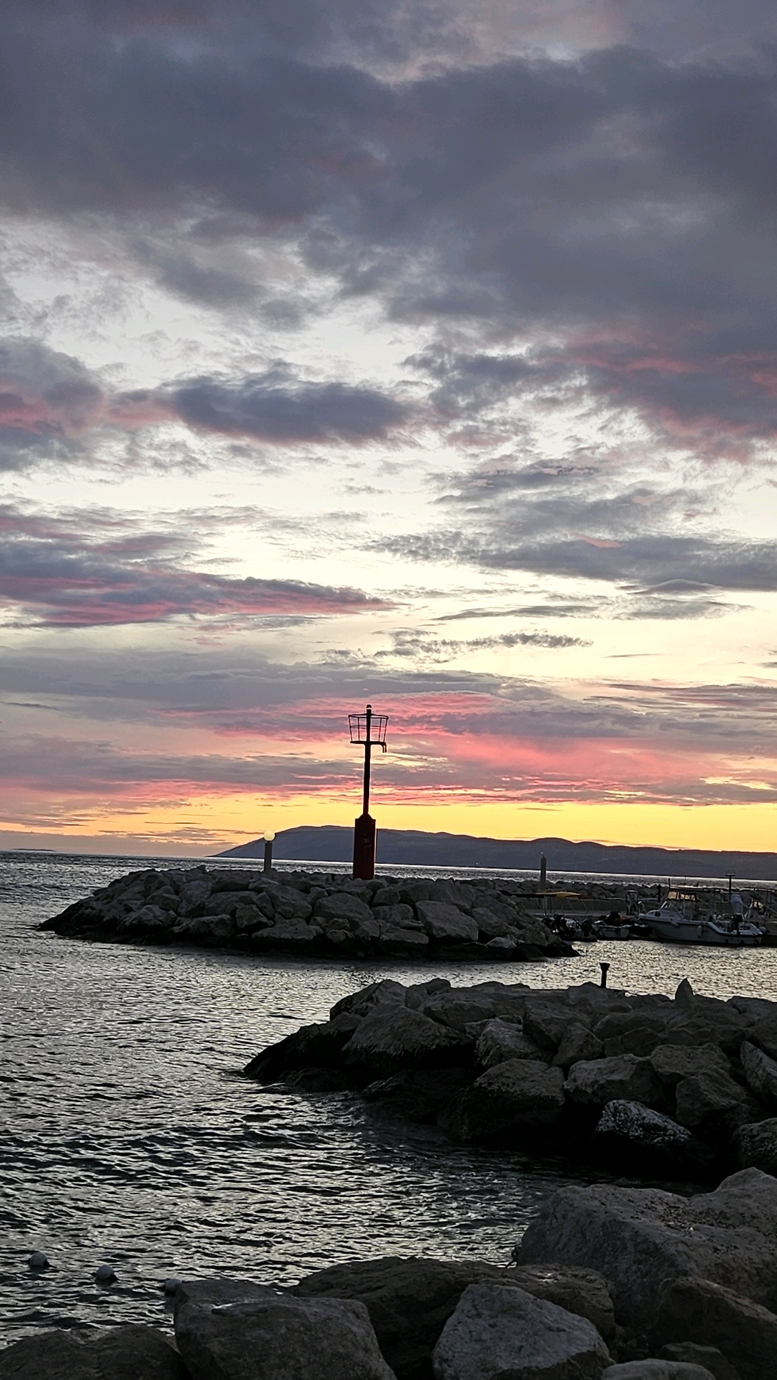 A vertical shot captures a sunset over the sea near Općina Tučepi, Croatia. The sky is painted with streaks of pink, orange, and gray clouds. In the foreground, a rocky breakwater extends into the water, topped with a red and black navigation beacon. The sea is calm, with small waves lapping against the rocks. In the distance, a hazy shoreline and mountains are visible. Several small boats are docked in a marina to the right. The lighting suggests the scene is captured during the golden hour before dusk.