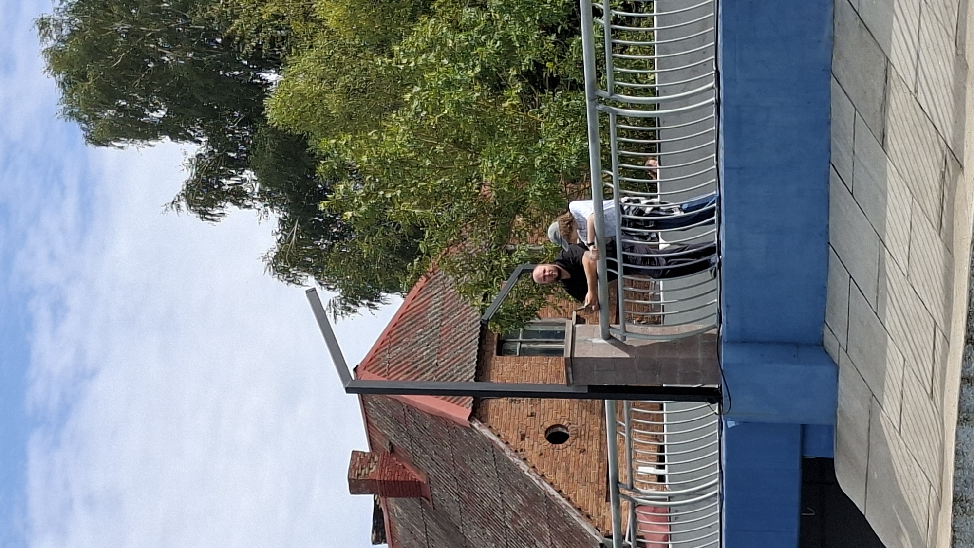 A man and a woman stand on a pedestrian bridge in Pastavy, Belarus, leaning on the railing. The man, with a shaved head and wearing a black t-shirt and dark pants, holds a smartphone in his left hand. The woman, wearing a grey cap, a white shirt with a pattern, and dark pants, stands next to him, looking in the same direction as him. Behind them, a red-tiled roof of a brick building with a chimney is visible, along with lush green trees under a bright sky with scattered clouds. The bridge has a metallic railing and a blue lower section. The ground leading up to the bridge is paved with large grey tiles.
