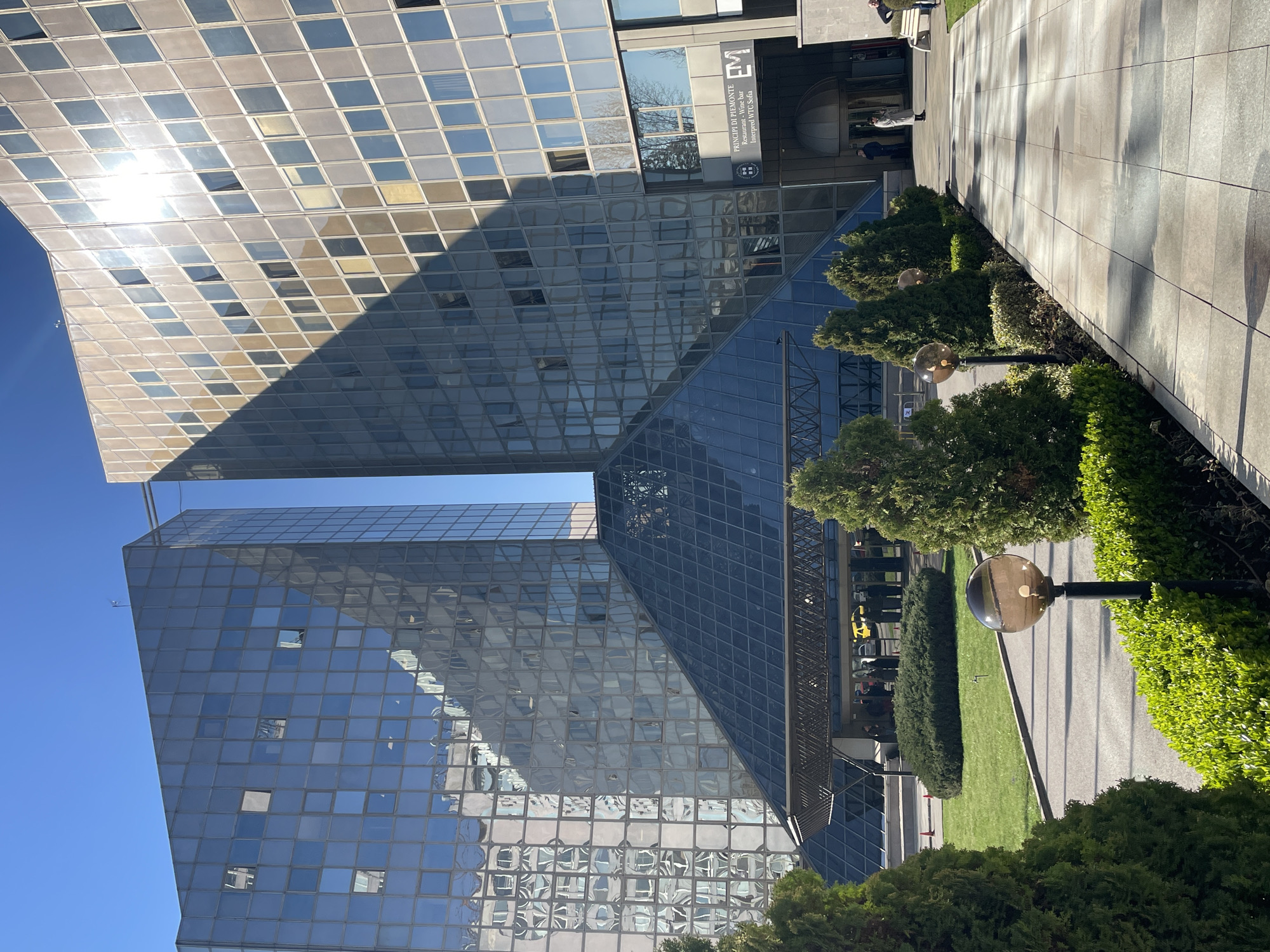 Here's a detailed description of the image:
**Overview:** The image shows a modern architectural scene with glass-facaded buildings, landscaping, and pedestrian areas.
**Location:** Based on the signage visible, specifically "Principi di Piemonte Ristorante - Wine-bar interpreted WTC Sofia," this is likely a location within or near the World Trade Center Sofia in Sofia, Bulgaria.
**Buildings:** The main focus is on a pair of modern buildings. Both have facades constructed from many square or rectangular glass panels, creating a grid-like appearance. The buildings' shapes are somewhat complex with slanted or pointed rooflines. A structure with a pyramid roof connects to the building.
**People:** There are several people visible in the background, appearing as small figures walking or standing near the building entrances.
**Landscaping:** Manicured greenery and landscaping are present along a walkway. This includes trimmed hedges, small trees, and grassy areas. A series of globe-shaped light fixtures on black poles lines the walkway.
**Walkway:** A tiled or paved walkway runs parallel to the building. The walkway appears to be in good condition, with some shadows falling across it.
**Environment:** The sky is clear and blue, suggesting a sunny day. Light is reflecting off the glass facade of the buildings.
