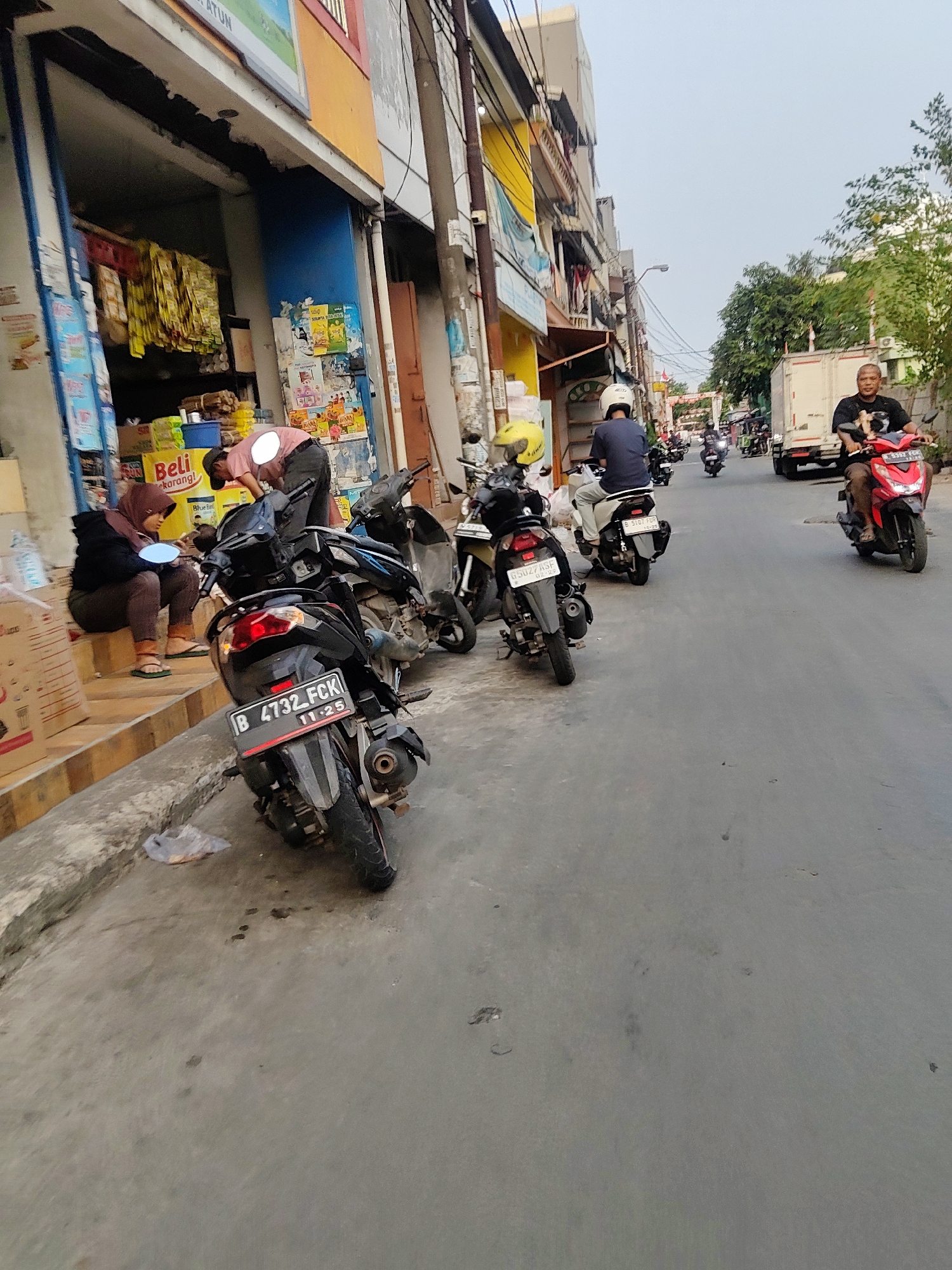 The image captures a street scene in Mekarmukti, Indonesia, depicting a narrow road lined with buildings on the left and a clear sky above. Several motorbikes are parked and in motion. In the foreground on the left, two people are sitting and standing near the entrance of a shop. A woman wearing a headscarf and dark clothing sits on the steps of the shop, while a person wearing a pink shirt and a cap is bent over near the shop entrance. A black motorbike with license plate B 4732 FCK is parked prominently in the foreground. Further down the street, other motorbikes are visible, with some riders wearing helmets. A white truck is parked on the right side of the road. The buildings on the left appear to be shops or residences, with various goods displayed outside one of them. The road surface is asphalt, and there are some scattered debris and oil stains on the ground. The overall impression is a typical urban street environment with moderate activity.