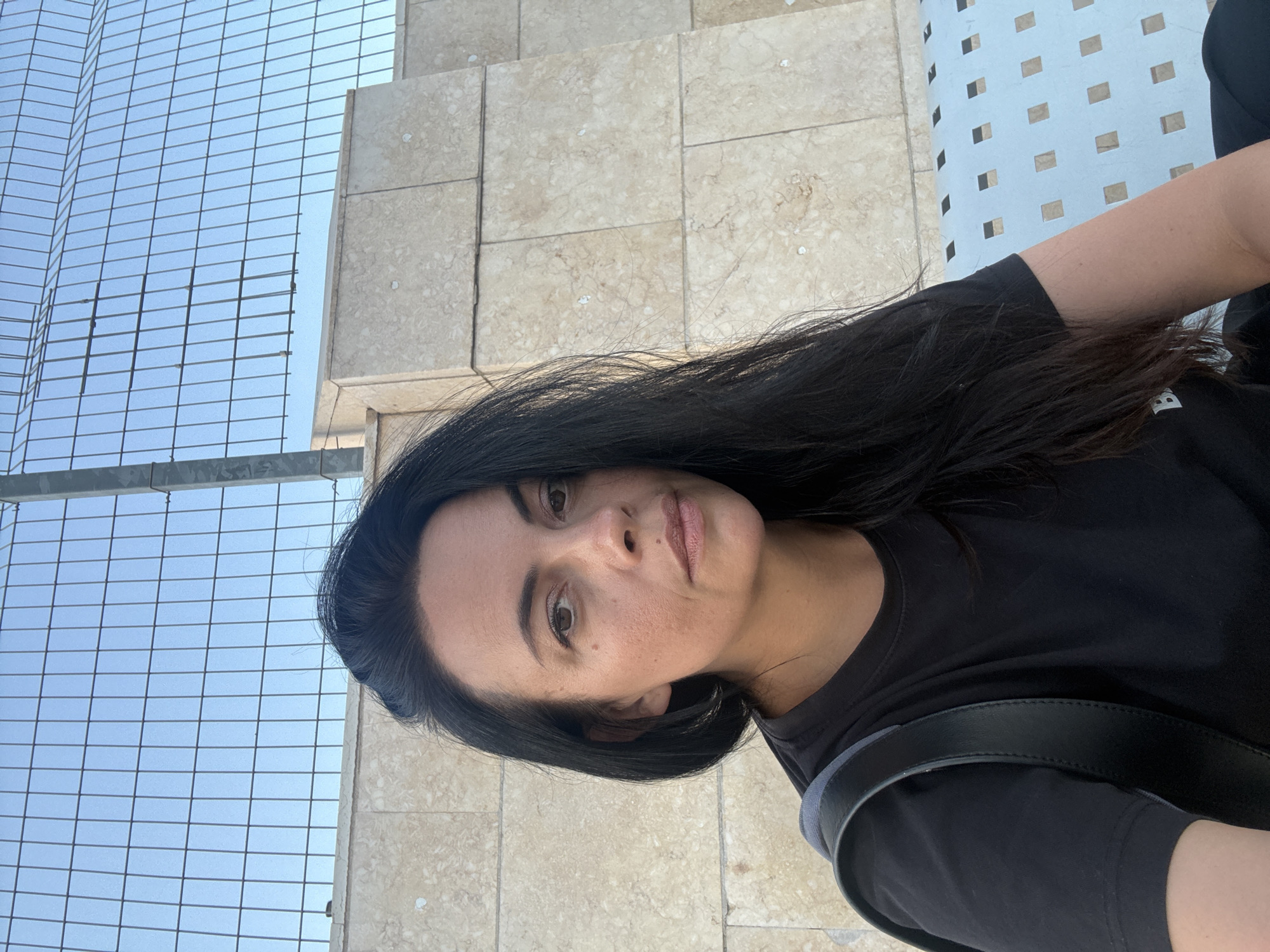 A woman with long, dark hair and fair skin is seated on a light blue bench with a square pattern. She is wearing a black t-shirt and has a black backpack strap visible over her left shoulder. The background consists of a light-colored, textured wall composed of large, square stone blocks, and a metal fence with a clear sky behind it. The overall setting appears to be an outdoor public space, potentially within the city of Hod HaSharon, Israel.